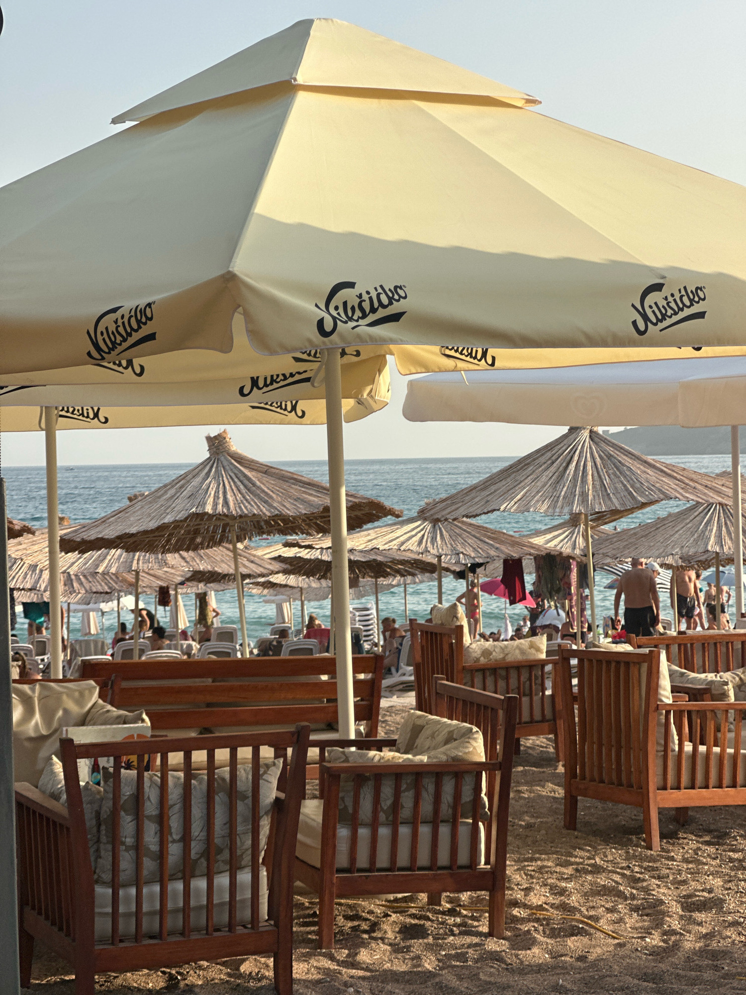 The image depicts a beach scene in Boreti, Montenegro, featuring a seating area on the sand with wooden chairs and tables under large, cream-colored umbrellas. The umbrellas have black lettering that reads "Nikšićko". In the background, the blue sea is visible, with people sunbathing on lounge chairs and walking along the shore. Several straw umbrellas provide shade on the beach. The lighting suggests it is daytime, with sunlight casting shadows. The overall atmosphere appears relaxed and leisurely.