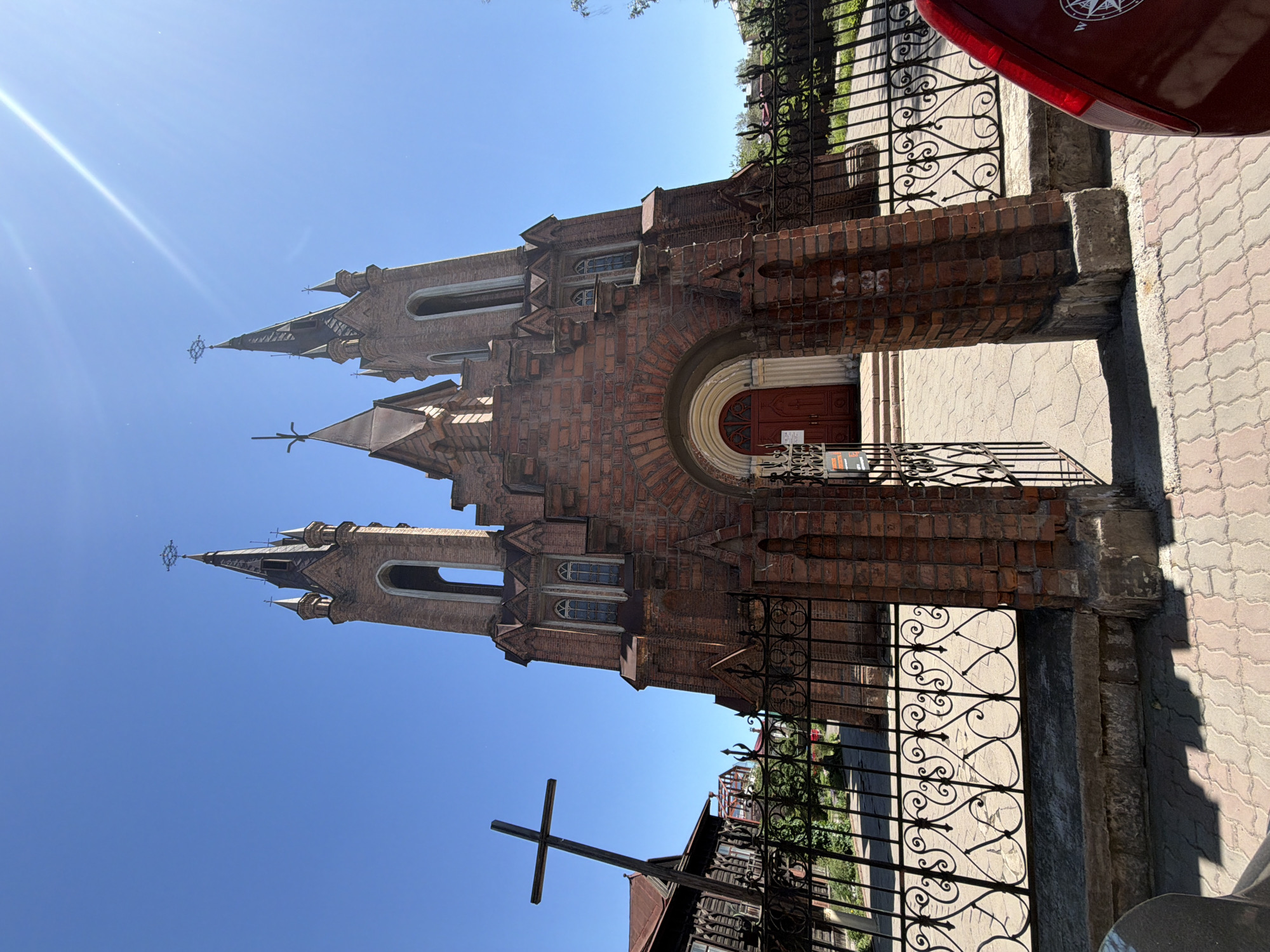 The image depicts the exterior of a red brick church with Gothic architectural elements, set against a clear blue sky. The church has two prominent towers with spires, a central arched entrance with red doors, and decorative window structures. In the foreground, there is an open black metal gate with ornate scrollwork, leading to a paved walkway. To the left of the church, a tall wooden cross stands tilted. The surrounding area appears to be a city, with hints of other buildings and greenery visible behind the fence. The image is taken from a low angle, emphasizing the grandeur of the church. The location is identified as the Catholic Church of the Holy Trinity in Krasnoyarsk, Russia.