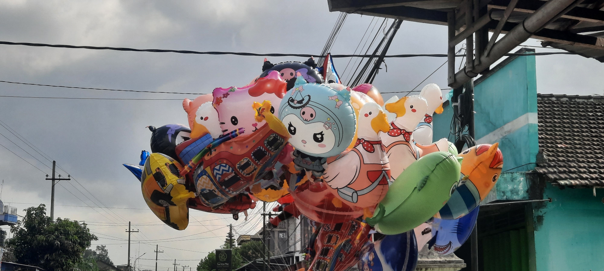 A cluster of inflated foil balloons, featuring cartoon characters like a pig, ducks, a cat, and a shark, are displayed outdoors. The balloons are attached to strings and appear to be suspended, possibly for sale. In the background, utility poles and power lines are visible against a cloudy sky. To the right, a turquoise building with a tiled roof and a wooden awning can be seen. The overall setting suggests an outdoor market or street vendor stall in a city, identified as Jatitengah, Indonesia.