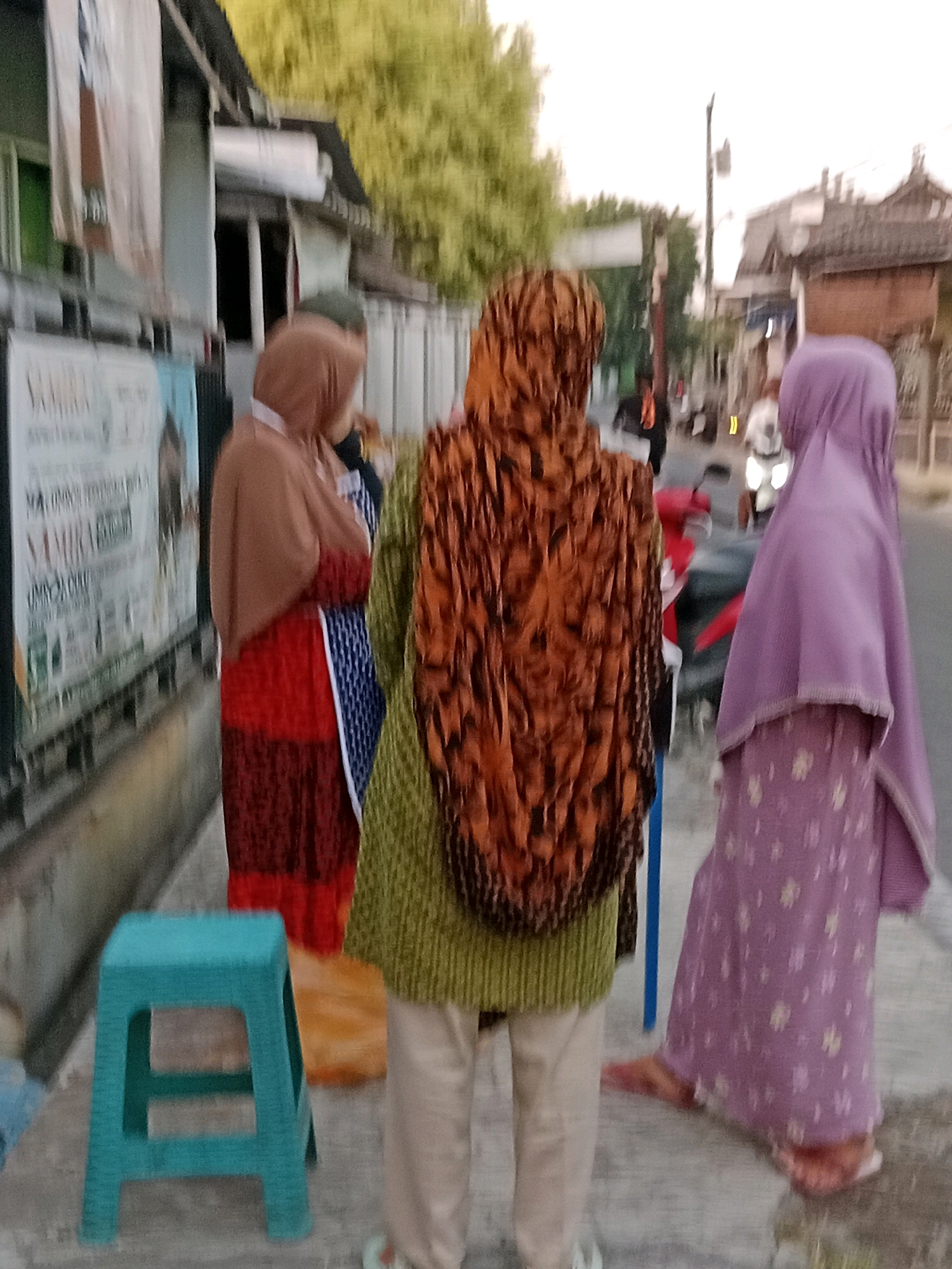 The image depicts an outdoor scene in Waru, Indonesia, possibly along a street or sidewalk. In the foreground, a teal plastic stool is visible on the right side of the frame. Several individuals, all appearing to be women wearing hijabs, are positioned in the mid-ground. The woman closest to the viewer, on the right, wears a light purple hijab and a long purple dress with a floral pattern. To her left, a woman in the center wears a green long-sleeved top and a headscarf with a prominent orange and brown patterned design. Her lower body is obscured by the frame. To the left of the center figure, another woman is visible wearing a brown hijab and a red and blue patterned dress. In the background, the environment suggests an urban or semi-urban setting with buildings, trees, and what appears to be a street with some vehicles, including a motorcycle. The overall lighting indicates it is likely daytime, possibly late afternoon or early evening. The image has a slightly blurry quality.