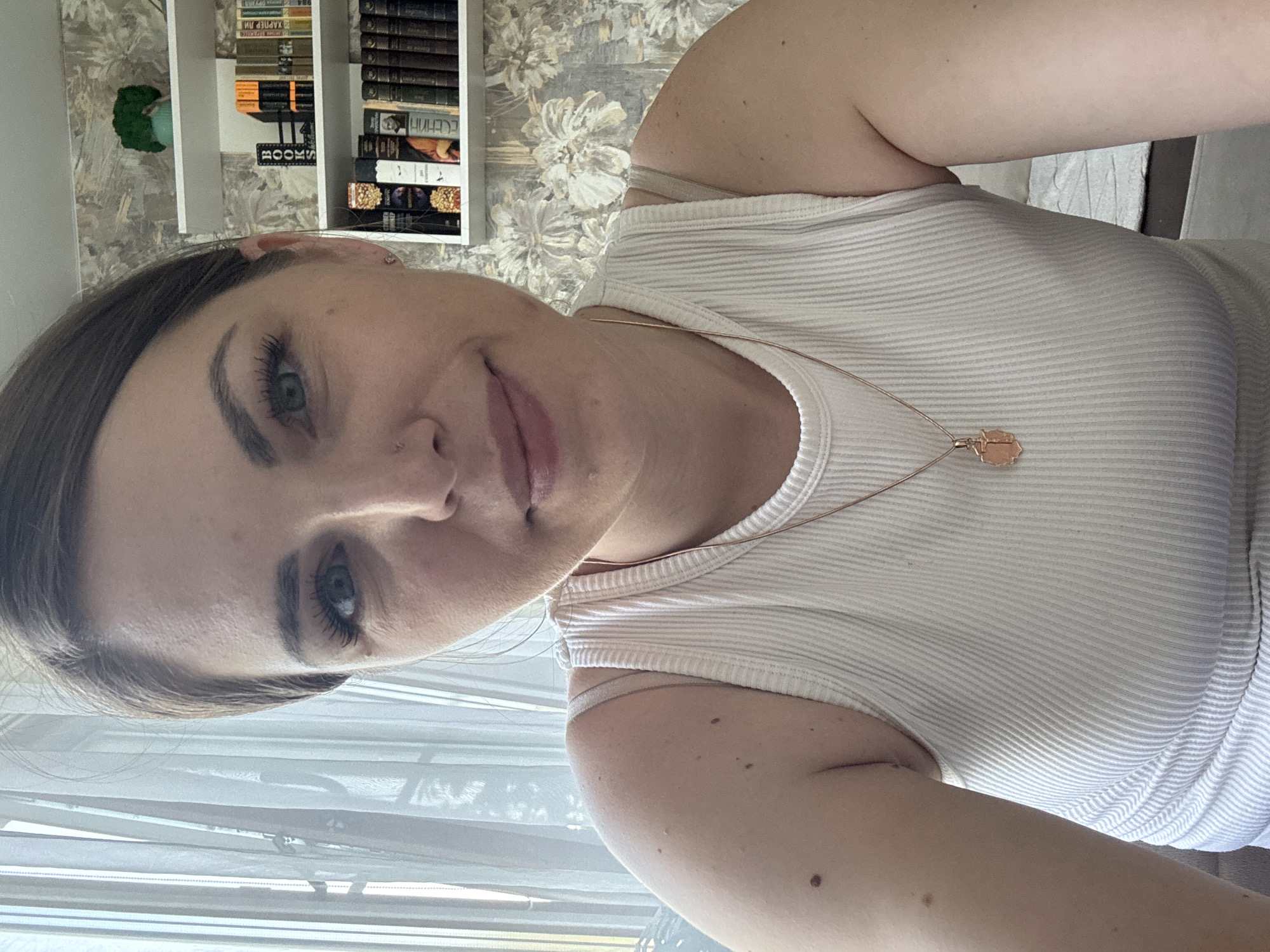 A woman is centered in the frame, wearing a white ribbed tank top and a delicate gold necklace with a pendant. She is facing the camera, slightly angled, with a neutral expression. Her hair is brown and pulled back from her face, and her eyes are light blue.
Behind her, slightly out of focus, is a bookshelf filled with books, and a patterned wallpaper is visible. Natural light is coming from a window to her left, which has sheer curtains.
The image does not indicate any information related to Razdolye, Russia.