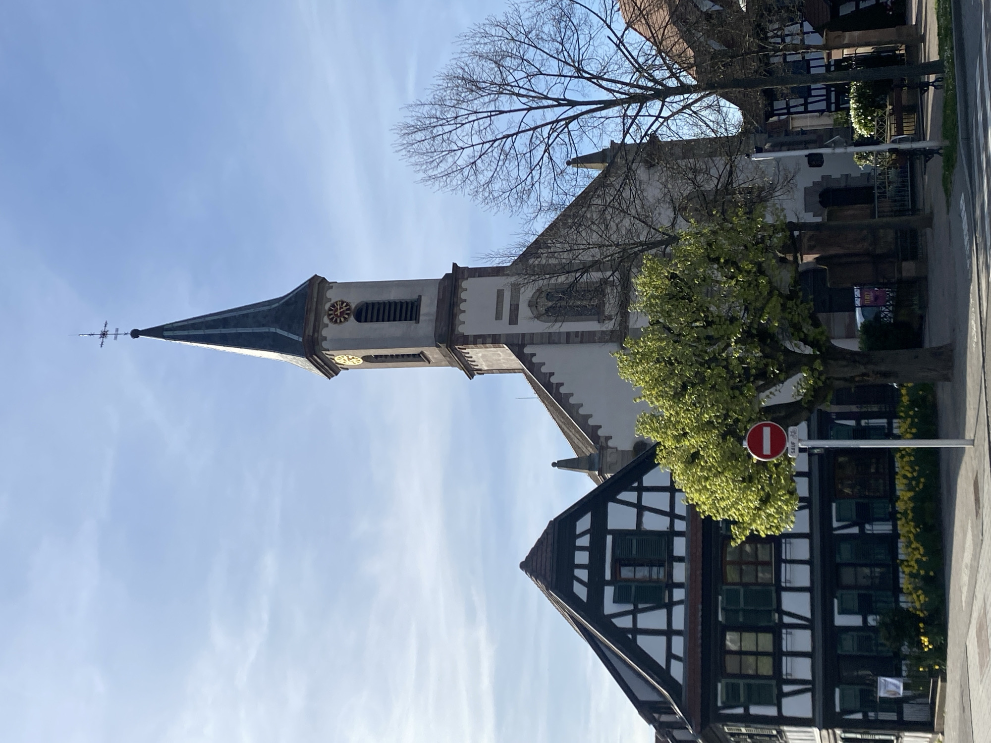 The image shows a view in Hoenheim, France, featuring the steeple of a church and a portion of a half-timbered house against a blue sky with light clouds. The church steeple is tall and narrow with a clock face and a pointed roof, topped with a weather vane. A bare tree is visible to the right of the church. Below, a corner of a traditional half-timbered house with dark wooden beams and white walls is seen. A tree with green leaves partially obscures the house. A "no entry" sign is also visible near the tree. The image is taken from a low angle.