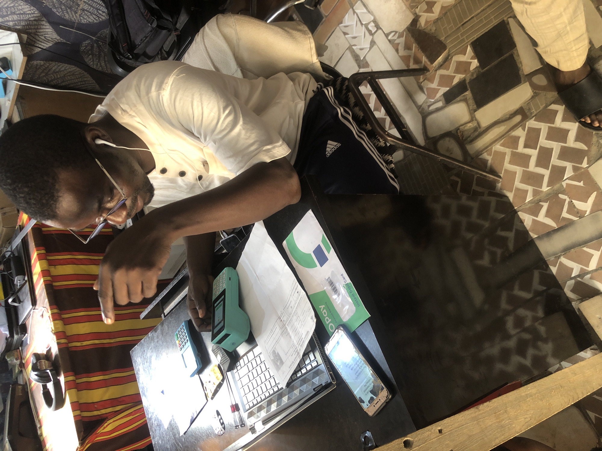 The image captures a medium shot of a Black man sitting at a desk in what appears to be a small office or shop in Ado, Nigeria. He is wearing glasses, a white short-sleeved collared shirt, and dark blue Adidas shorts. The man has earbuds in and is pointing his right index finger towards the viewer, as if in discussion or explanation. On the desk in front of him are a laptop, two point-of-sale devices (one blue, one teal), a smartphone, and some paperwork. The desk surface is dark and reflective, showing the patterned floor tiles beneath. Behind the desk, a striped cloth is visible, possibly a table runner or backdrop. The overall impression is of a person engaged in business activities, likely related to financial transactions given the presence of the POS devices and the OPay branded item. The lighting suggests it is daytime, with shadows cast by the man and objects on the desk. The floor is tiled with a mosaic pattern of brown and beige squares.
