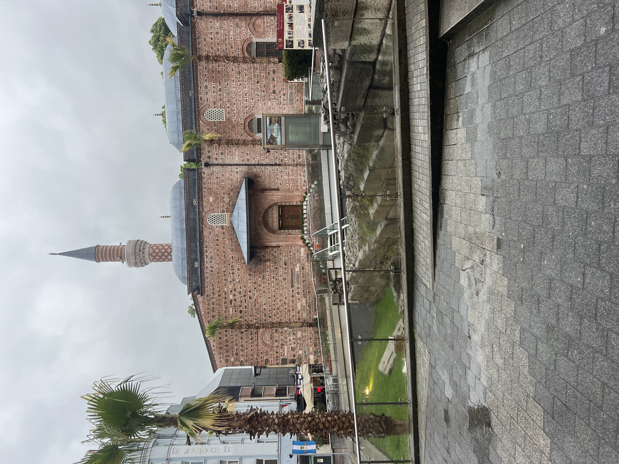 The image captures a street scene in Plovdiv, Bulgaria, featuring a large historic building, likely a mosque, dominating the left side. The building is constructed with alternating courses of red brick and grey stone, featuring multiple grey domes topped with finials and arched windows, some of which have decorative grilles. A tall, slender minaret, built with similar brick and stone patterns and capped with a pointed spire and a balcony, stands prominently to the left of the main structure.
In the foreground and extending to the right, there's a grey tiled paved area, appearing damp as if from recent rain. A modern glass railing with metal supports runs along this paved area, overlooking a lower section with green grass and what appear to be ancient stone foundations or archaeological ruins.
Two large palm trees with robust trunks and green fronds are visible in the foreground, to the bottom left. Modern buildings with glass facades and signs are partially visible behind these trees and in the background. The sky is overcast.
Notable details on the historic building include a large informational panel with unreadable text and images, and below it, a digital display showing pictures. The overall scene blends historical architecture with contemporary urban infrastructure. No specific activities or people are visible.