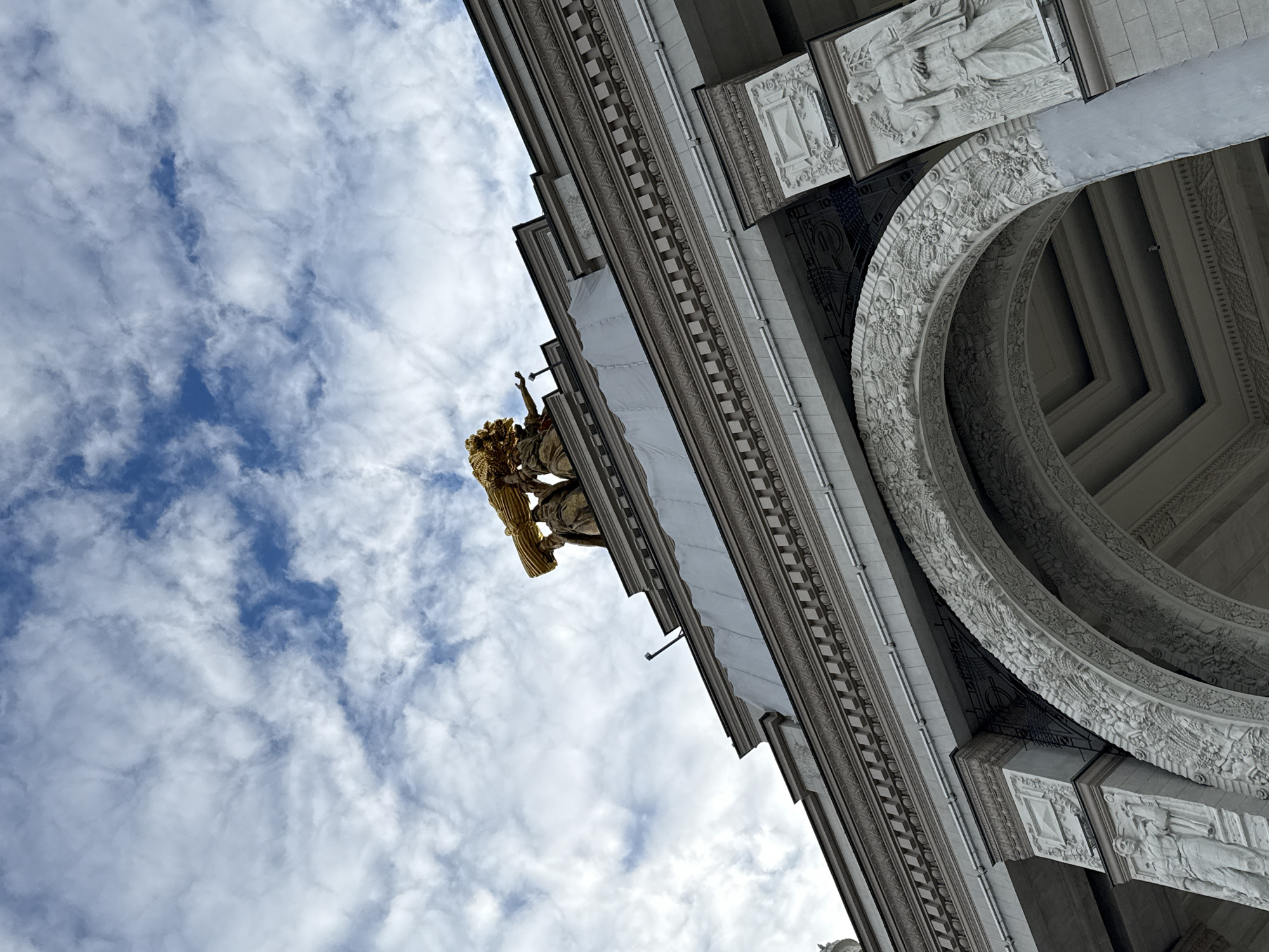 The image presents a dramatic low-angle perspective of a grand, ornate architectural structure, likely a triumphal arch or monumental building, viewed from below against a bright, partly cloudy sky. The architecture is primarily composed of light-colored, possibly white or grey, stone or concrete, showcasing rich classical and neoclassical detailing.
A large, curved archway dominates the right side of the frame, its inner and outer surfaces extensively decorated with intricate relief carvings. These carvings depict a variety of elements, including figures, garlands, floral motifs, and geometric patterns, creating a sense of historical grandeur. Above and to the left of the arch, the building's facade rises, featuring multiple horizontal bands with detailed ornamentation such as checkerboard patterns and scrolling foliage. Interspersed within these bands are rectangular relief panels, one clearly showing a classical figure in robes holding a staff or implement, further emphasizing the monument's historical or allegorical themes.
Prominently featured towards the upper left is a striking golden sculptural group, perched on a cornice or ledge. This element, partly obscured, appears to be a section of a larger composition, possibly a quadriga or a group of allegorical figures, with bright golden accents catching the light.
The sky is a mix of scattered white cumulus clouds and clear blue patches, indicating a bright daytime setting, possibly morning or late afternoon given the soft, diffuse lighting. No specific human activity or event is depicted, and no visible text is present. The monumental scale, classical design, and the prominent golden sculpture are strong indicators of a significant landmark, consistent with structures found in Moscow, Russia, such as the Triumphal Arch.