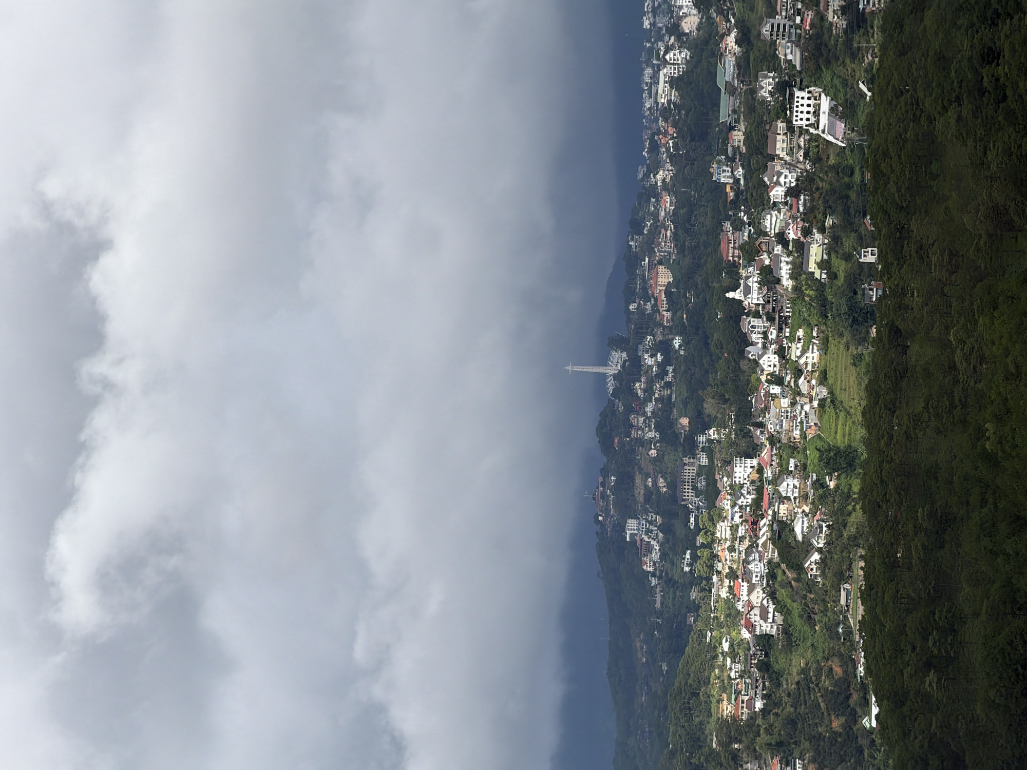 The image is a high-angle shot of a densely populated town nestled within rolling green hills under a heavy, overcast sky. Numerous houses with a variety of architectural styles and roof colors are scattered across the hillsides, some with manicured gardens. A prominent white structure resembling a statue with outstretched arms stands tall on a distant hilltop. In the foreground, a thick band of dark green pine trees forms a dense forest. The overall atmosphere is moody and somewhat subdued due to the cloudy weather. The location is the city of Xuân Thành, Vietnam.