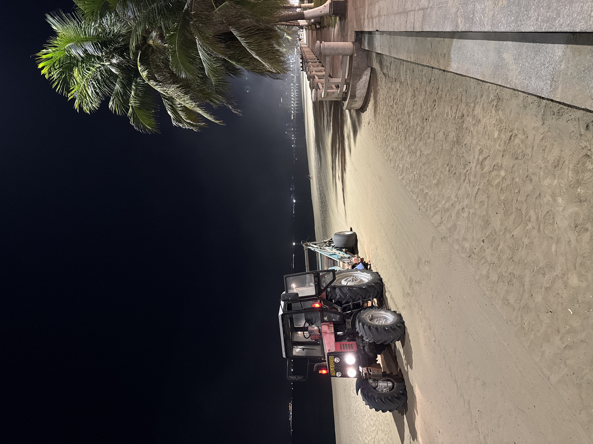 A red tractor with a trailer is parked on a sandy beach at night in Nha Trang, Vietnam. The tractor's headlights are on, illuminating the sand. The beach is next to the sea, with palm trees and a walkway lining the shore. In the distance, city lights and buildings are visible. The sky is dark, and the scene is lit by artificial lights.