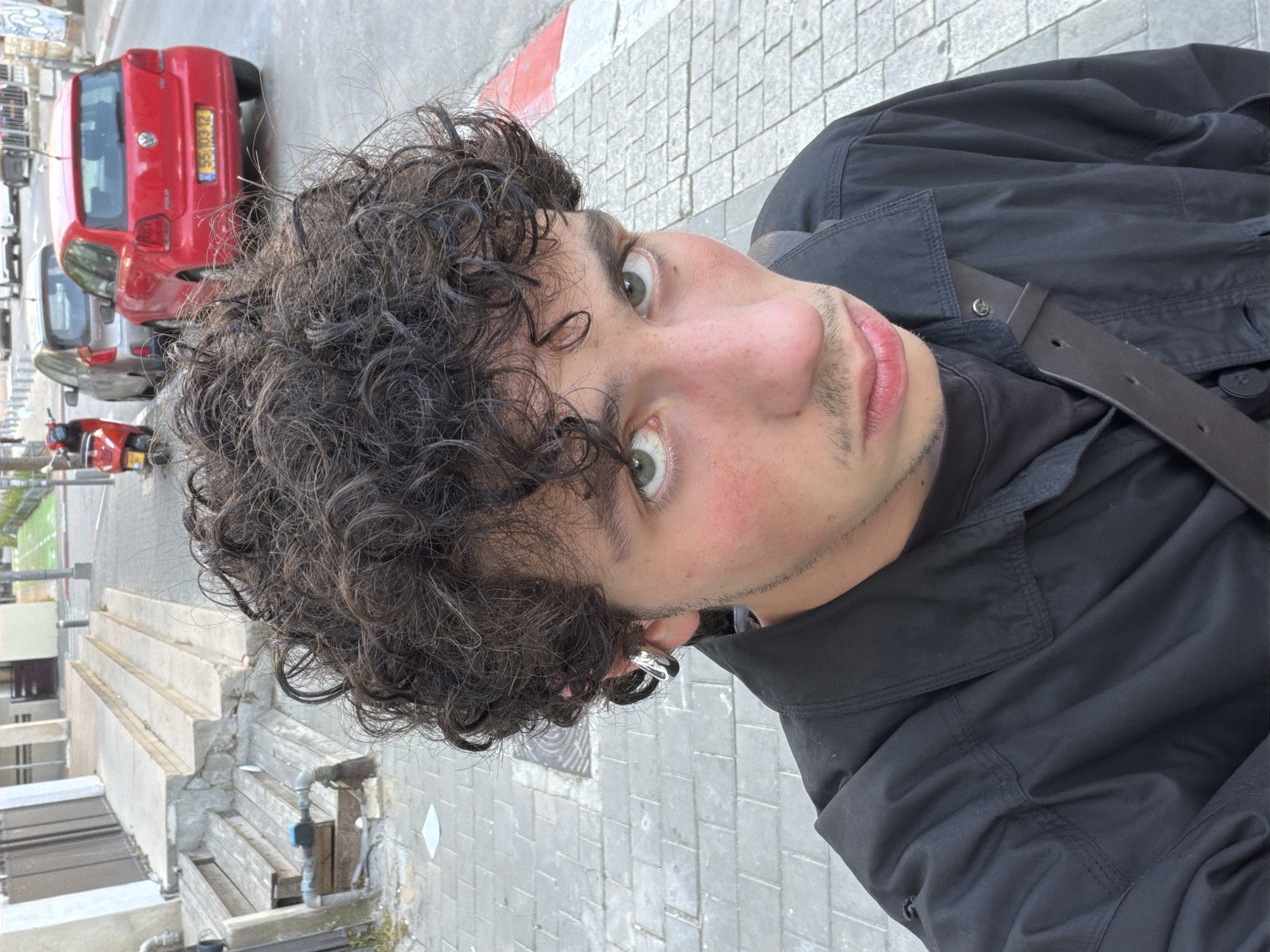 A medium close-up shot features a young man with curly brown hair, green eyes, and fair skin. He is wearing a black jacket and a silver hoop earring. A dark brown belt crosses his chest. The background shows a sidewalk with gray tiles, and in the distance, red and gray cars are visible. Further in the background, there is a staircase structure and greenery. Based on the vehicles' license plate, it is most likely set in Tel-Aviv, Israel.