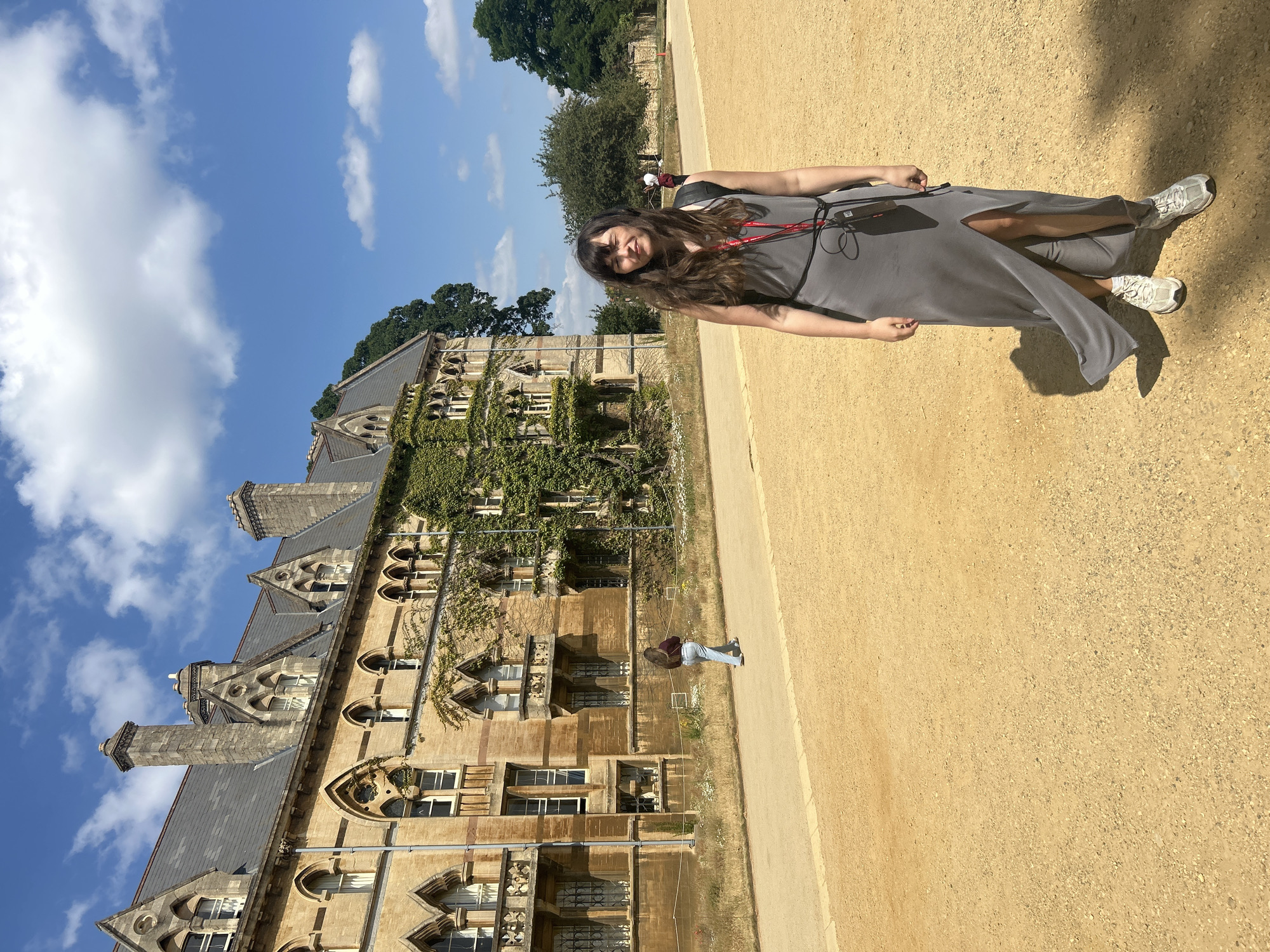 An outdoor, medium shot captures a woman standing on a gravel pathway in front of a large, historic stone building with gothic architectural elements, possibly in Oxford, United Kingdom. The woman, with long brown hair and bangs, wears a long, grey dress with a high slit and a black belt, and white sneakers. A black backpack and a lanyard with a red strap are visible around her neck. She stands with her eyes closed and a slight smile.
Behind her, the building displays arched windows, stone carvings, and is partially covered with ivy. The sky above is blue with scattered white clouds. A person in jeans and a burgundy shirt walks away from the camera on the pathway in the background. The pathway is a light brown gravel, and there are patches of dry grass and a few small white flowers along the side of the building. The overall lighting suggests a sunny day.