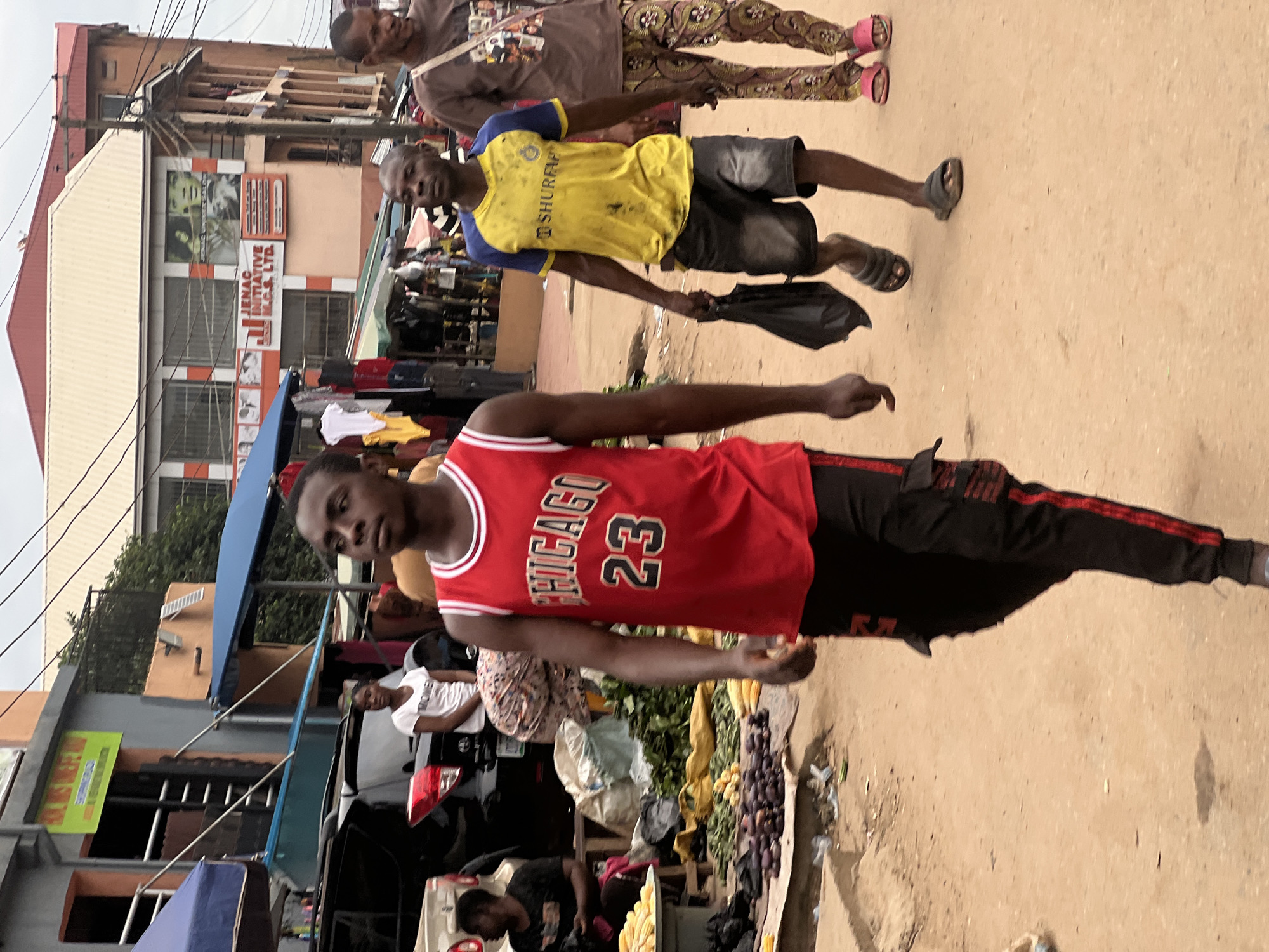 A medium shot captures three men walking on a dirt road in Nkpor, Nigeria, with a busy market scene in the background. The man in the foreground, on the left, wears a red sleeveless basketball jersey with "CHICAGO 23" printed on it, and black pants with red stripes. He is walking away from the camera, looking forward. To his right, a man in a yellow and blue t-shirt with the logo "SHURE" is walking and holding a black plastic bag. He wears dark shorts and dark sandals. On the far right, a man in a brown t-shirt and patterned trousers walks with pink sandals on his feet. Behind them, a market stall displays various items including produce and clothing. A woman in a white t-shirt is visible in the background near a black SUV. The environment is dusty and appears to be a busy urban area with buildings and power lines visible. The sky is overcast.