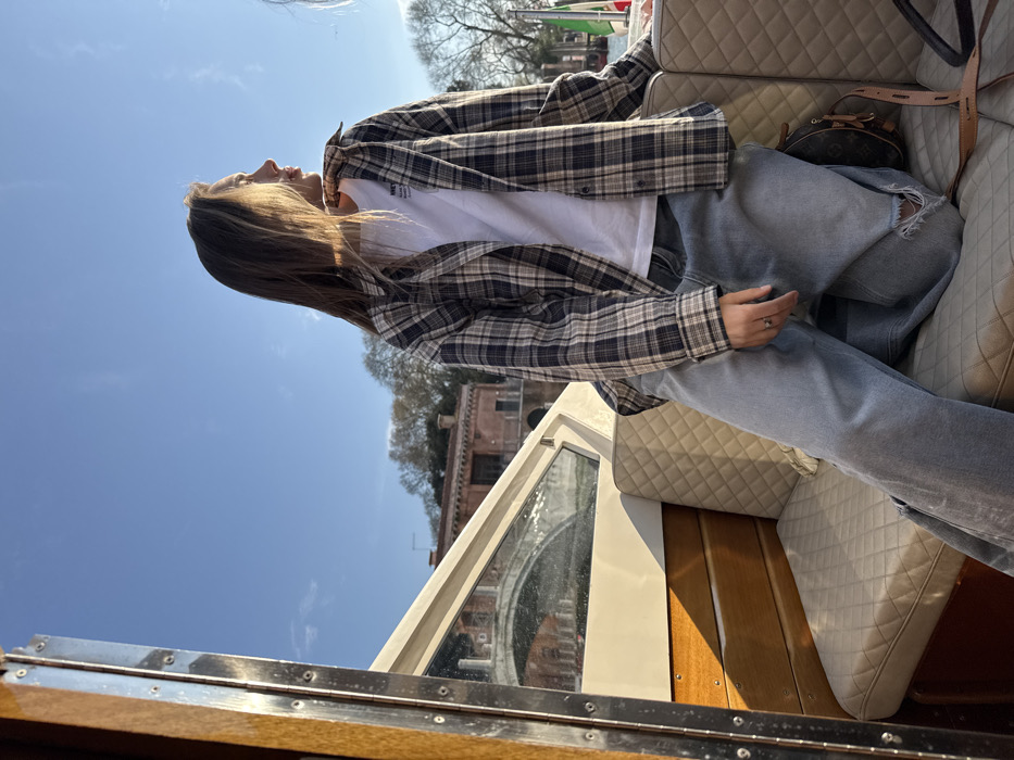 The image depicts a young female seated in a boat on a canal in Venice, Italy, under a clear blue sky. The female, with long brown hair illuminated by sunlight, wears a white t-shirt underneath an unbuttoned plaid shirt in shades of dark blue, beige, and white, paired with light blue distressed jeans featuring a tear on the right knee. She is seated on a cream-colored, diamond-quilted upholstered bench seat, looking towards the right side of the frame. Her left hand rests on her left thigh, displaying a ring on the ring finger. A brown monogrammed bag with a leather strap is visible on the seat beside her right leg. The boat's interior features light wooden paneling and a metal-trimmed window structure on the left. In the background, beyond the boat's structure, traditional Venetian architecture is visible, including a reddish-brown building and a light-colored stone arch bridge spanning the canal. Sparse trees are present on the distant bank, and a partial view of an Italian flag (green, white, and red vertical stripes) is visible on the far right. The scene is brightly lit by strong sunlight.