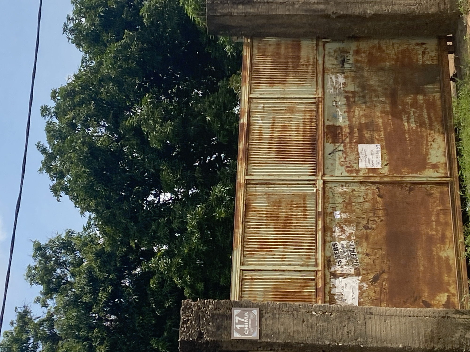 The image shows a rusty metal gate in front of dense green trees, with a blue sky visible above. On the left side of the gate, a concrete pillar has a sign that reads "17 CHIMA STREET". The gate itself is old and weathered, with several posters and papers attached to it. The overall scene suggests an outdoor environment in a city, likely Ado, Nigeria, given the text on the sign. The gate appears to be a barrier to entry, and the posters may be advertisements or notices. The presence of lush trees indicates a possibly tropical or sub-tropical climate.