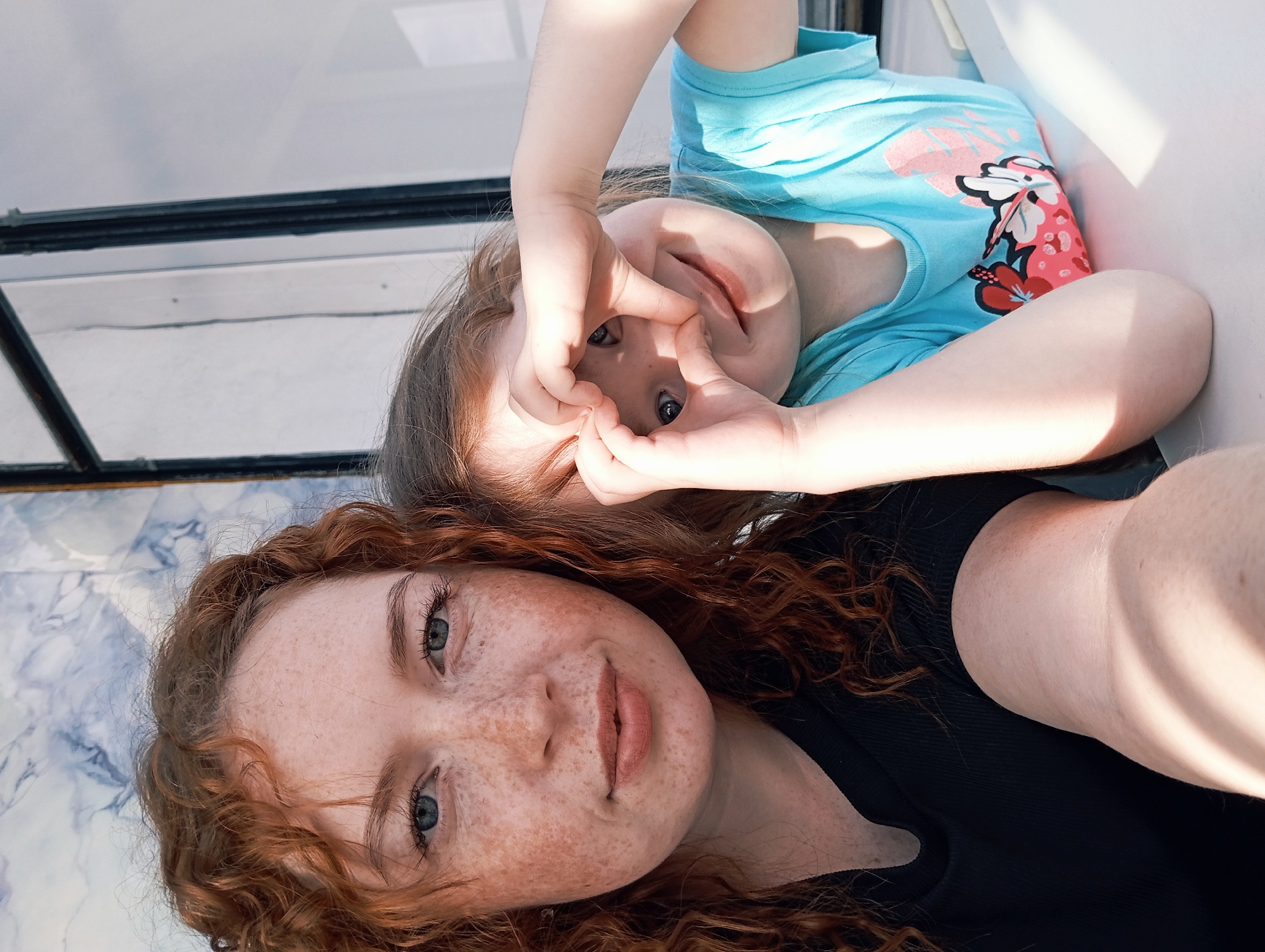 This brightly lit image, rotated 90 degrees clockwise, captures a close-up selfie of two individuals, likely a mother and child, in a casual setting. The adult, with vibrant red, curly hair, an abundance of freckles across her nose and cheeks, and light blue eyes, occupies the foreground. She gazes directly at the camera with a soft, content expression, wearing a dark top.
Behind her, and appearing upside down in the rotated frame, is a child with light brown hair and blue eyes. The child playfully frames her eyes with her hands, creating a heart-like shape, and looks through this opening towards the viewer. She is dressed in a light blue t-shirt featuring a distinctive pink and red tropical or floral print.
The background includes a dark-framed window or glass panel, suggesting an indoor or enclosed space. A light-colored surface with a blue and white marbled pattern is visible near the adult's head. Strong, direct sunlight illuminates the scene, casting prominent highlights and shadows, indicating a sunny daytime environment. Both subjects convey a sense of calm engagement and playfulness. There is no visible text in the image.