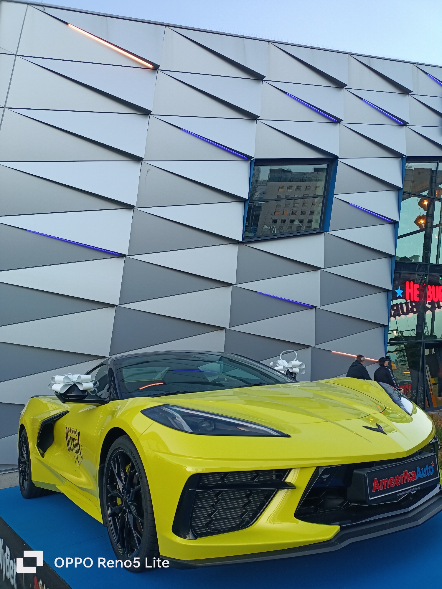 A bright yellow Chevrolet Corvette is displayed on a blue platform in front of a modern building with a geometric facade. The building's facade is composed of triangular panels in shades of grey and white, with embedded LED lighting in orange and blue. A reflection of an office building is visible in a window on the facade. Two people are standing near the building entrance, which has a red neon sign that reads "HE BUR". A license plate on the car reads "Ameriika Auto" with a website address. The bottom left corner of the image shows the text "OPPO Reno5 Lite". The scene appears to be an outdoor event or dealership in Tallinn, Estonia.
