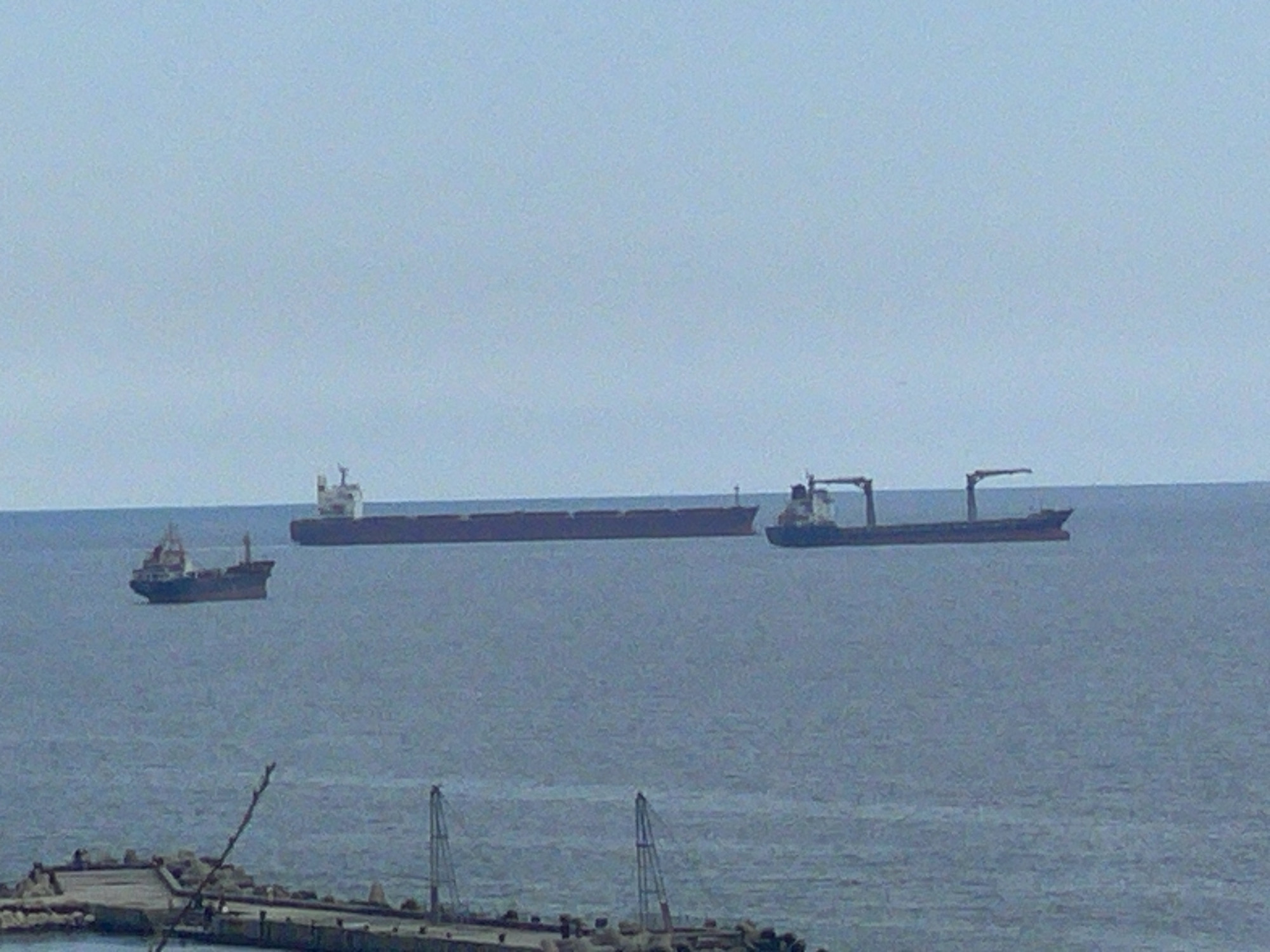 The image shows three cargo ships on the Black Sea near Varna, Bulgaria. A small cargo ship is to the left of the frame, with a large cargo ship in the middle, and a cargo ship with a crane on the right of the frame. In the foreground there is a breakwater or pier with several small gantry cranes. The sky is overcast.