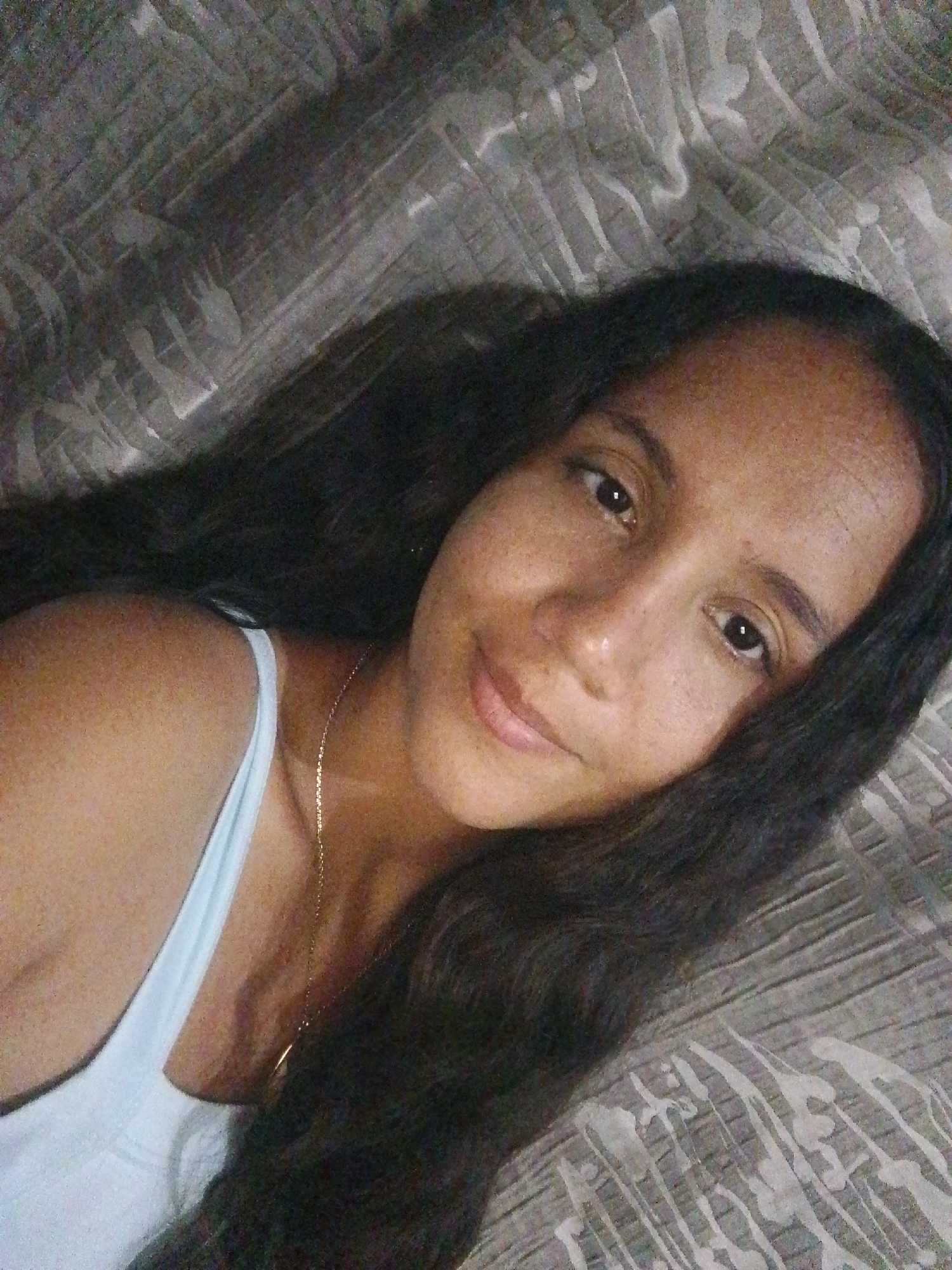 A woman with long, dark hair is lying down on a patterned bedspread. She is wearing a light blue tank top and a gold necklace. The background is a gray patterned fabric. The location is unknown; there is no indication this image was taken in Juan Griego, Venezuela.