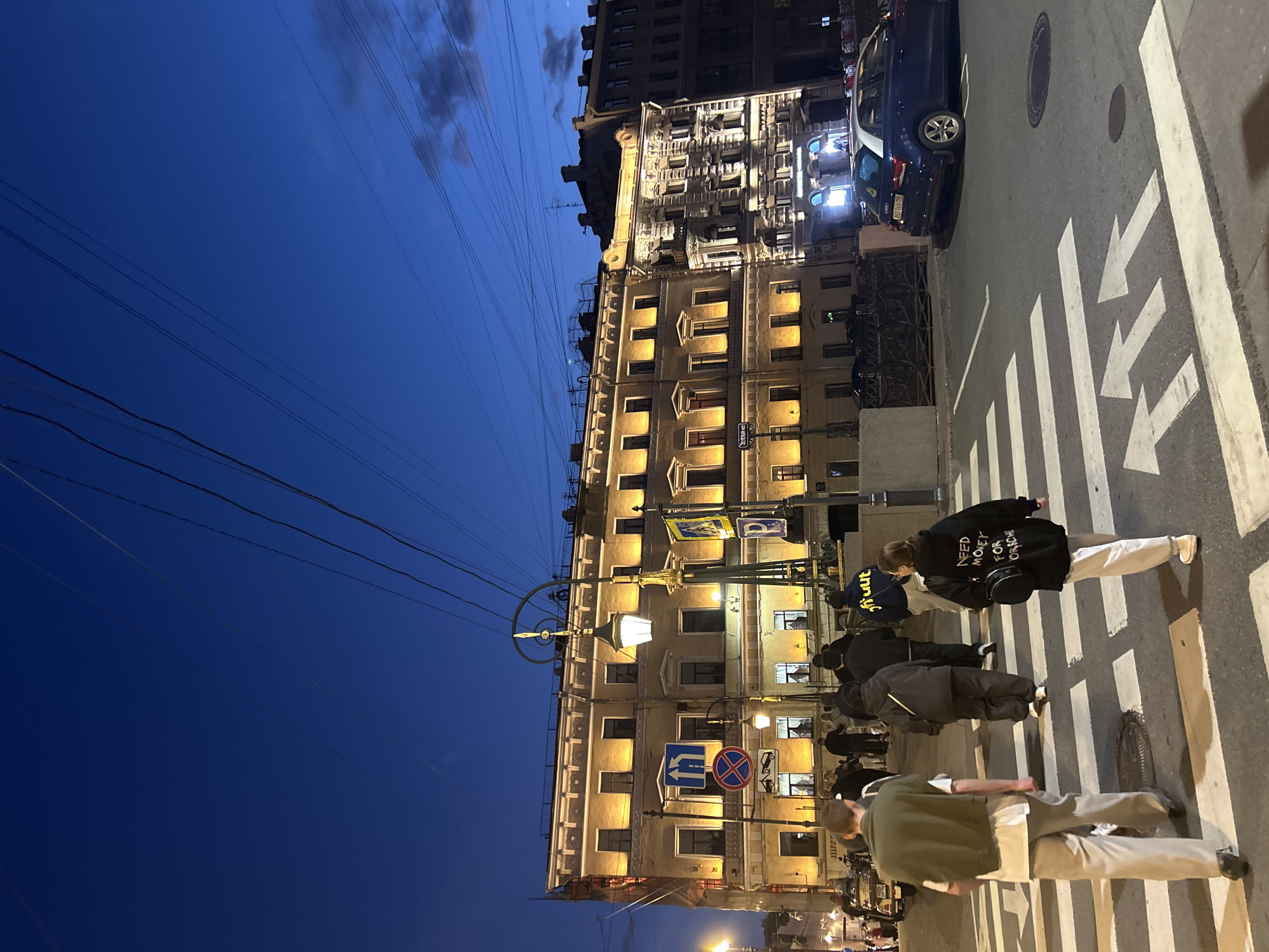 The image depicts an urban street scene at dusk, with a clear blue sky fading into darkness. A pedestrian crossing with white stripes and directional arrows is prominent in the foreground. Several individuals are crossing the street. A young woman in the foreground right wears a black hoodie with white text, carrying a black backpack. A young man in the foreground left wears a khaki jacket over a white t-shirt and light-colored pants. Behind them, a group of people are also crossing.
The background features a multi-story building illuminated with warm yellow lights, showcasing architectural details like windows and cornices. Streetlights, one ornate and tall with a glowing lamp, stand near the building. Traffic signs, including a "no stopping" sign and directional signs, are visible on poles. A blue car is parked on the right side of the street, with its license plate partially visible. Overhead, numerous electrical wires crisscross the sky. The overall scene suggests a city environment in the evening.