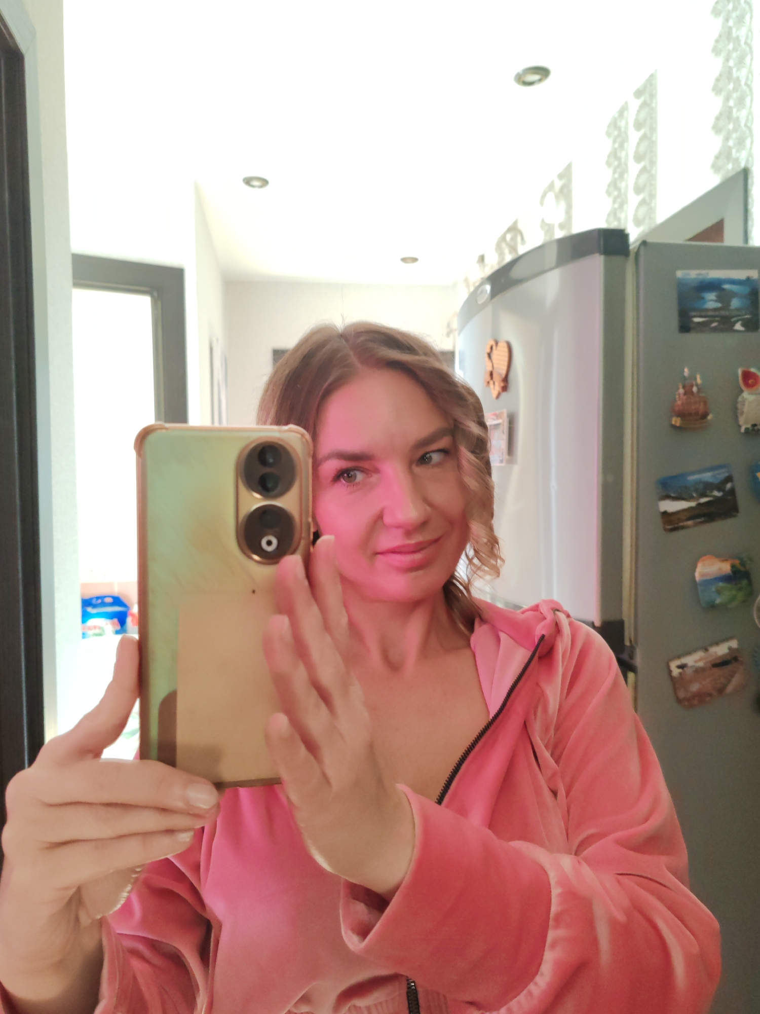 A woman is taking a selfie in what appears to be her kitchen. The woman has wavy brown hair and is wearing a pink tracksuit jacket. She is holding a gold-colored smartphone up in front of her. The kitchen background includes a gray refrigerator covered with magnets and other decorations. There's a doorway visible behind her with a light-colored room beyond. The ceiling has recessed lighting. The image does not provide specific evidence to determine if the image was taken in Petropavlovsk-Kamchatsky, Russia.