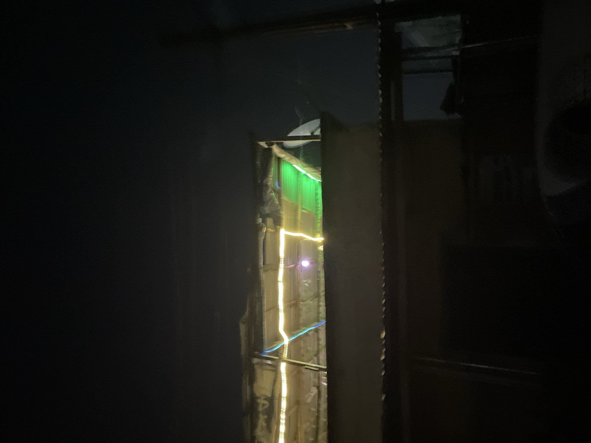 The image presents a dimly lit, claustrophobic interior scene, possibly a dwelling in Ado, Nigeria. A narrow opening or window reveals an exterior space illuminated by string lights and green neon tubing. The structure around the opening appears to be rudimentary, perhaps constructed from corrugated metal or similar materials. The foreground is dominated by dark, undefined shapes, hinting at structural elements of the building. The overall atmosphere is one of urban poverty, with makeshift construction and limited lighting. No people are visible.