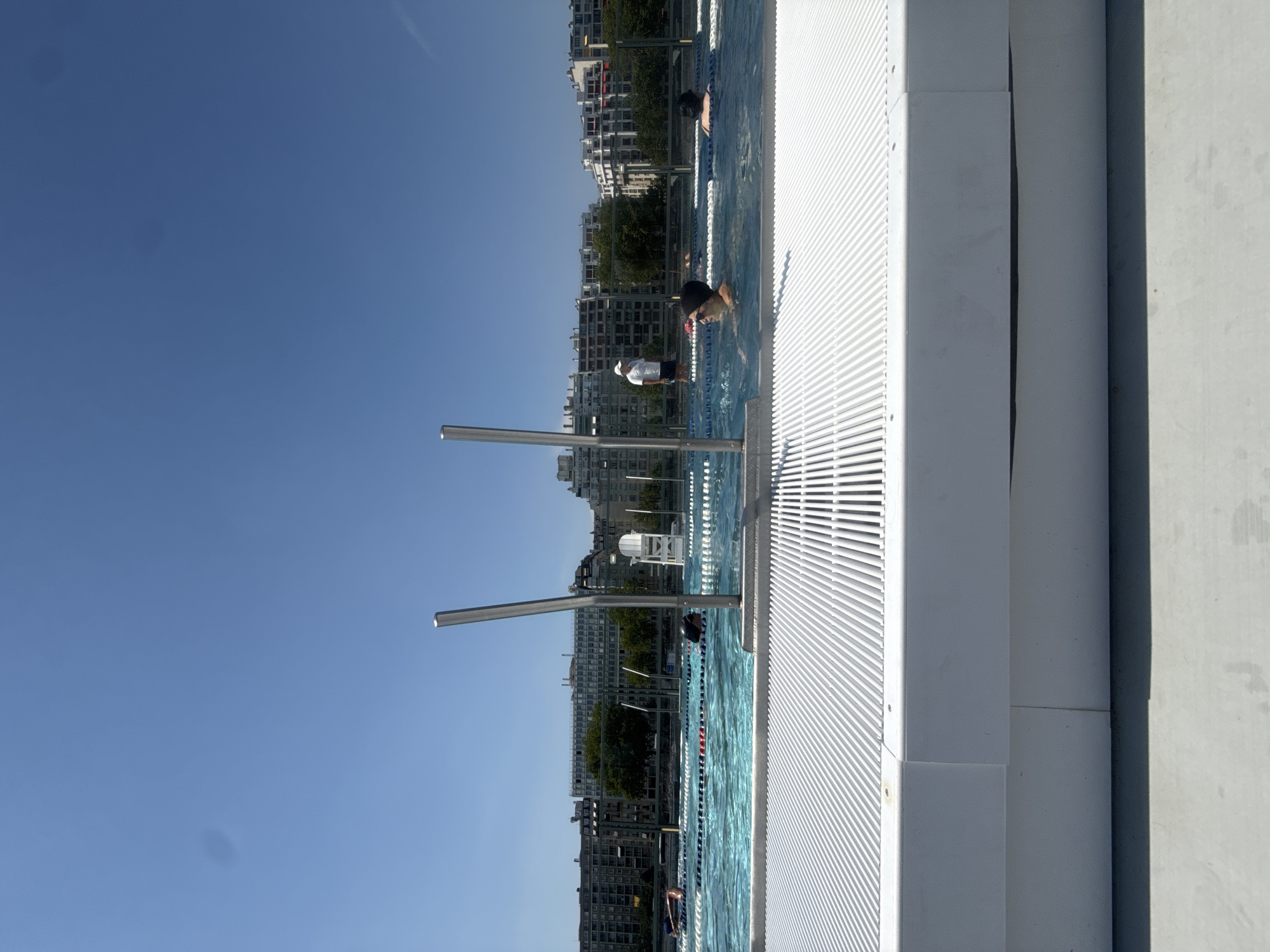 This media file depicts a swimming pool scene on a sunny day in Paris, France. The camera is positioned at an angle, looking down towards the water and the surrounding urban landscape. The foreground is dominated by the white, slatted edge of the pool, with two silver poles rising vertically from it, likely for support or to hold lane ropes.
In the pool, several people are visible. One person in the middle distance, wearing a white cap and a blue swimming suit, is standing in the water, looking towards the camera. Further down the pool, another person with a dark swim cap and goggles is swimming. In the far distance, a few more figures can be seen in the water. The pool is divided by lane ropes, indicating it's likely a public or training facility.
Beyond the pool, a dense urban backdrop rises, characterized by multi-story buildings with light-colored facades. Trees are interspersed among the buildings, suggesting a park or green space within the city. The sky is a clear, vibrant blue, indicative of good weather and likely midday. The overall impression is one of a pleasant, active day at a city swimming pool. No visible text is present in the media file.