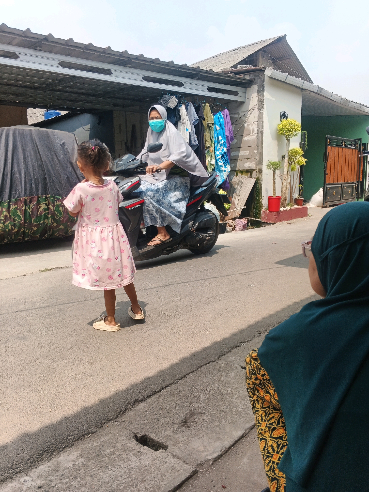 In Kadu, Indonesia, on a sunlit street, a young girl in a pink, Hello Kitty-patterned dress stands facing away from the camera. She wears light-colored slip-on shoes. Behind her, a woman wearing a light gray hijab and a teal face mask rides a black Honda motorcycle. The woman's lower body is draped in a blue and white patterned skirt, and she wears flip-flops. The motorcycle is parked on the right side of the road. In the background, a building with a corrugated metal roof is visible, with clothes hanging on a line in front of it. A dark-colored car cover is on the left side of the image. To the right, a green wall and a wooden gate are present, along with decorative potted plants. In the foreground, the back of another person, wearing a dark teal hijab and a patterned yellow and black skirt, is visible, looking towards the street scene. The street is paved with asphalt.