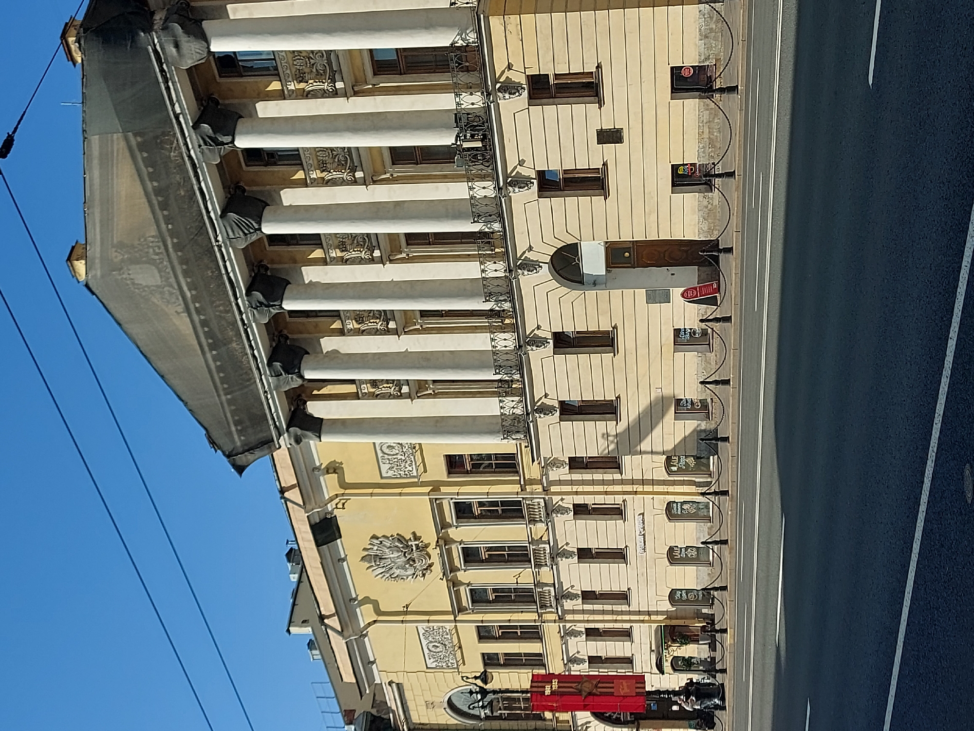 The image depicts a building with a classical facade in Saint Petersburg, Russia. The building is beige with white columns and decorative elements. It is located on a street with asphalt pavement and painted lane markings. A barrier of posts connected by chains runs along the building’s facade. Several commercial signs are attached to the building, indicating the presence of businesses. A section of the building is under construction or renovation, with a dark fabric cover. Overhead power lines are visible against a clear blue sky.