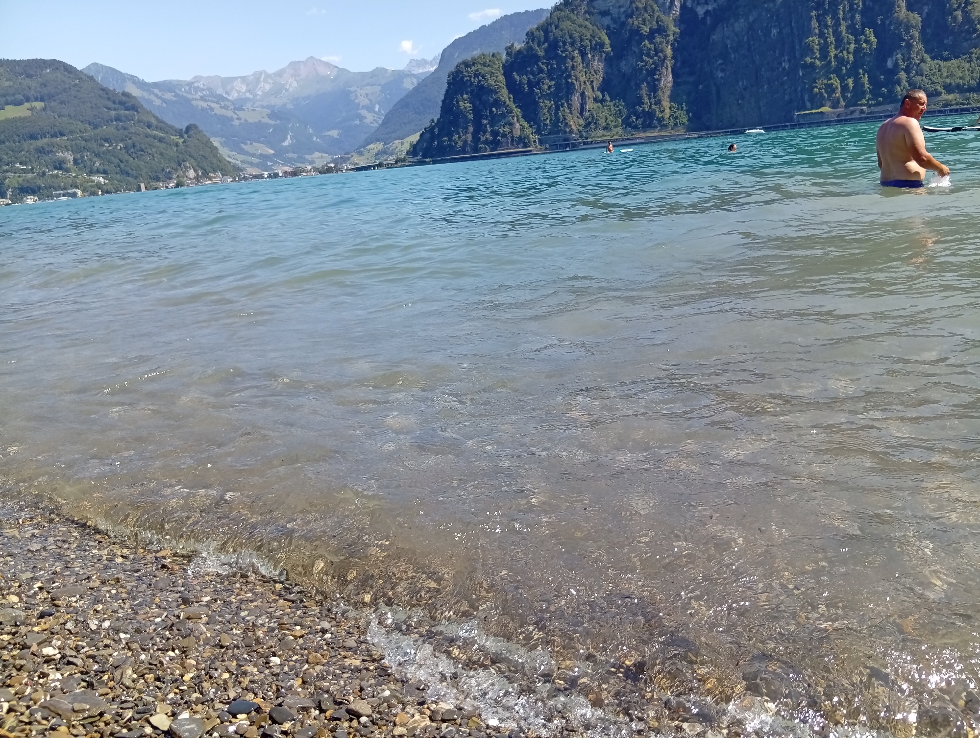 A man wearing blue swim trunks is partially submerged in the clear, blue water of a lake, with a rocky shore in the foreground. He is facing away from the camera and towards the right side of the frame, with his right arm raised and splashing water. In the background, large, green, tree-covered mountains rise steeply from the opposite shore of the lake. A road and some buildings are visible along the base of the mountains, suggesting an urban or semi-urban area. The sky is clear and blue. The overall scene depicts a sunny day at a scenic lake location, likely in Switzerland given the mountainous terrain.