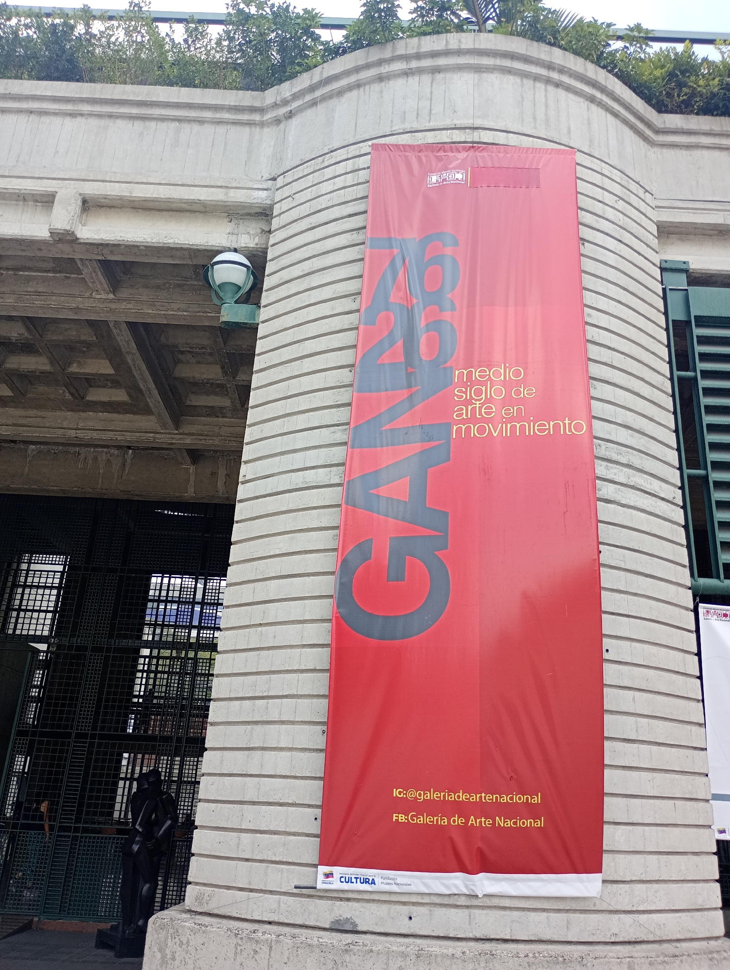 This daytime image showcases the exterior of a building, likely the Galería de Arte Nacional (National Art Gallery) in Caracas, Venezuela. A large red banner, prominently displayed on a curved, textured concrete wall, advertises an art exhibition. The banner features the bold text "GAND 26" and "medio siglo de arte en movimiento" (half a century of art in movement). It also displays social media handles: "IG:@galeriadeartenacional" and "FB: Galería de Arte Nacional," along with logos for "Fundación Museos Nacionales," "CULTURA," and "Venezuela," indicating a national cultural institution.
Above the banner, the concrete building features a decorative, curved frieze topped with green foliage. To the left, a heavy concrete overhead structure, possibly a bridge or an extension of the building, is visible with a distinctive waffle-slab ceiling. A white and green outdoor lamp fixture is attached to this structure. Below, dark metal grates or fencing enclose an area where a dark, abstract, human-like sculpture stands. A faint figure of a person's head and shoulder can be seen behind the grates on the far left. The scene suggests a public space, highlighting architectural elements and an ongoing cultural event. The weather appears clear with no strong shadows.