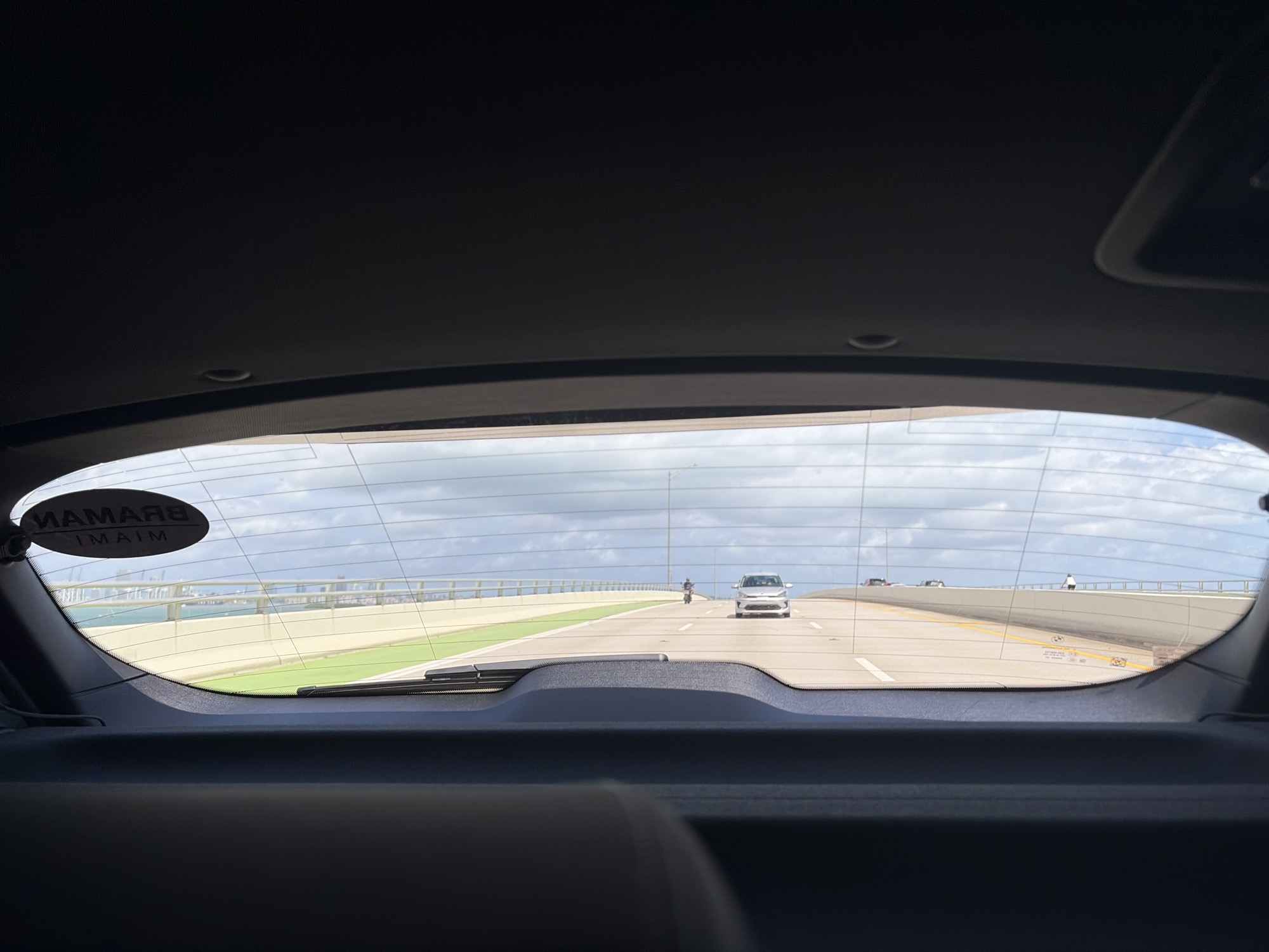 The photograph, taken from inside a vehicle, shows a view through the rear windshield. The vehicle is traveling on a bridge. Outside, there is a green space alongside the road, with a body of water visible in the distance. There are white barriers on the right side of the frame and an overpass can be seen through the back windshield. In the distance, there are other vehicles, a motorcyclist and a pedestrian visible. There is a sticker on the top-left corner of the windshield that says "Braman Miami", suggesting that the location is in Miami, United States of America. The sky is overcast with many clouds.