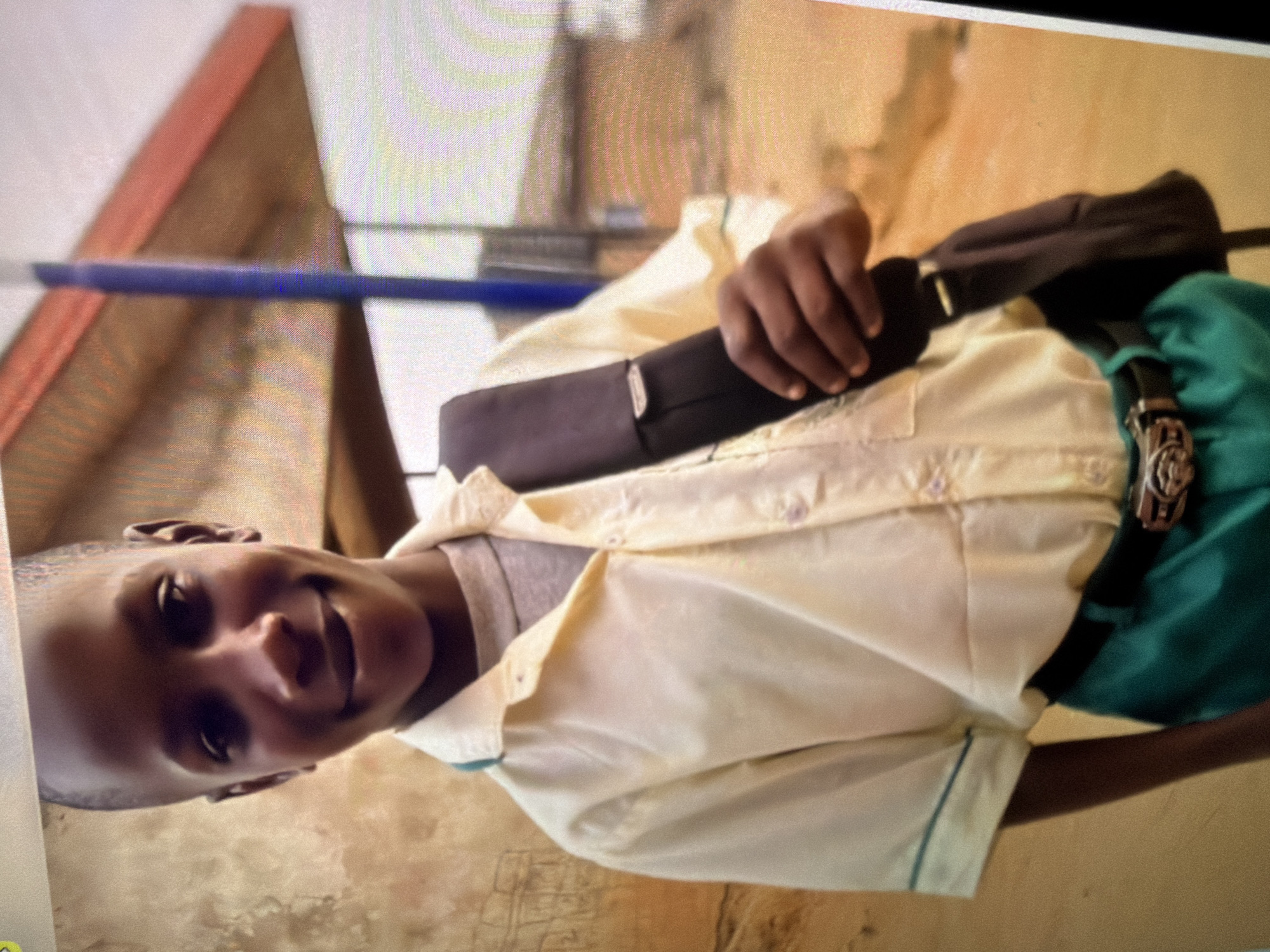 A young Black male is standing and smiling at the camera in an outdoor setting. He is wearing a light yellow collared shirt with a gray t-shirt underneath, and bright green pants with a black belt. He has a black backpack slung over his right shoulder, and is holding the strap with his right hand. The background is out of focus but appears to be a building with a reddish-orange roof, and a dirt path. There is a blue pole in the background. The overall lighting suggests it is daytime.