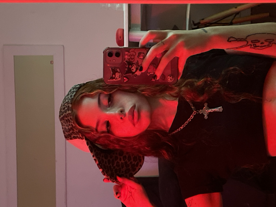 A young female individual is depicted in a mirror selfie, illuminated by a dominant red light source. She possesses long, wavy, auburn hair with lighter highlights. She wears a leopard-print baseball cap backward, adjusting it with her left hand, which has dark-painted fingernails. Her face, partially obscured by the phone, features a septum piercing and dark eye makeup. She is wearing a black, short-sleeved crop top and a silver-colored chain necklace with a crucifix pendant. On her inner left forearm, a linear black-ink tattoo of a stylized figure is visible. Her right hand holds a dark-colored mobile phone with a protective case adorned with multiple cartoon characters, prominently featuring two rear camera lenses. The background, a reflection of an indoor space, shows a light-colored rectangular frame on a wall and a white shelf with a small red object to the right. A wooden object, possibly a tripod or furniture, is partially visible in the lower right. The exact location within the city of Buenos Aires, Argentina, cannot be determined from the provided image.