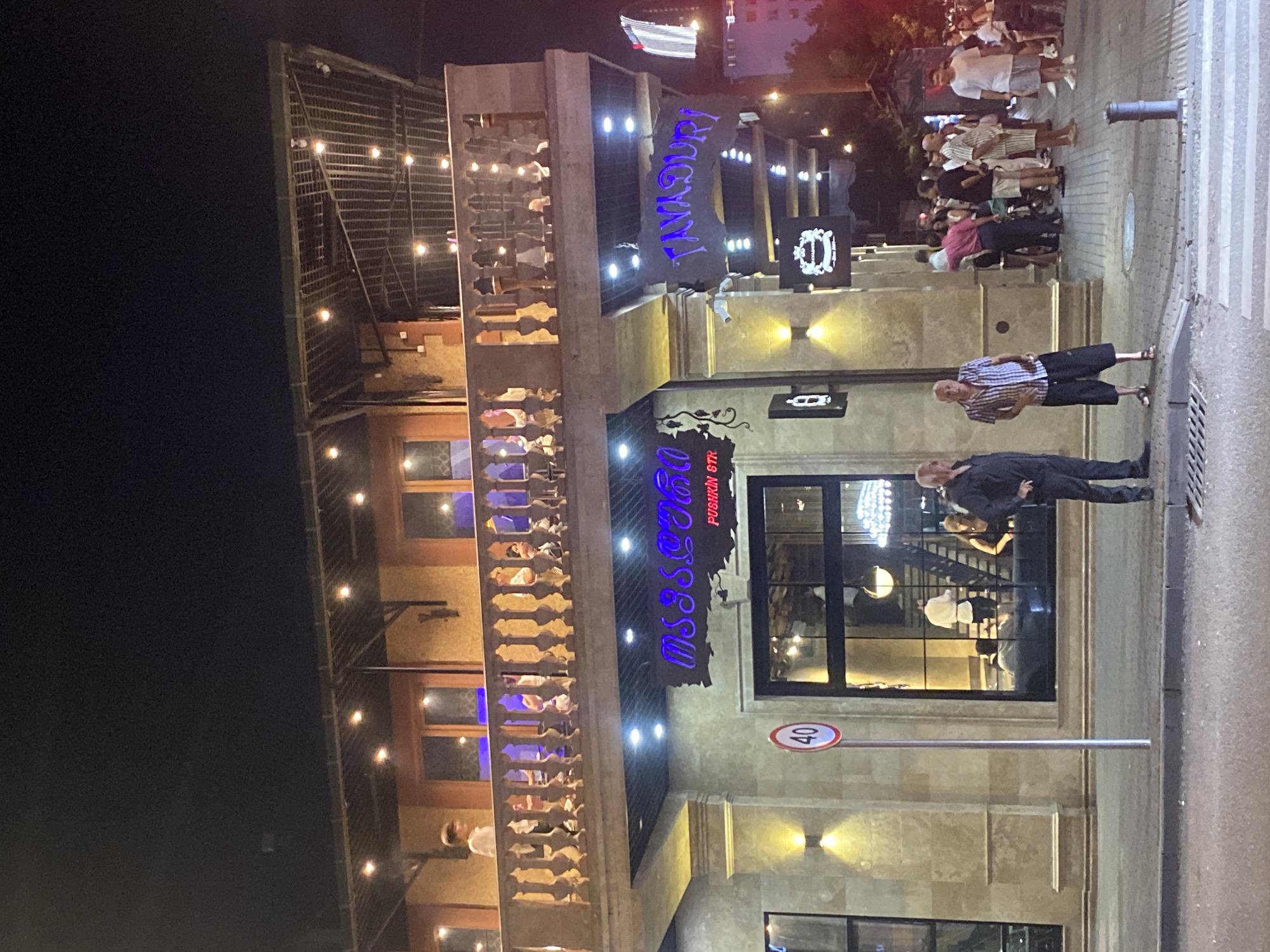 This is a night-time, eye-level, medium shot of the exterior of a multi-story establishment in Batumi, Georgia, likely a restaurant or bar. The building has a prominent second-story balcony with a decorative railing, illuminated by string lights. On the ground floor, neon signs in a foreign script, possibly Georgian, are visible. One sign reads "TAYADURI" in blue neon letters. A traffic sign indicating a 40 km/h speed limit stands near the entrance. Several people are gathered on the sidewalk in front of the building, some appearing to be waiting. The street in the foreground is paved, with a pedestrian crossing visible. The overall atmosphere suggests a lively urban setting at night.