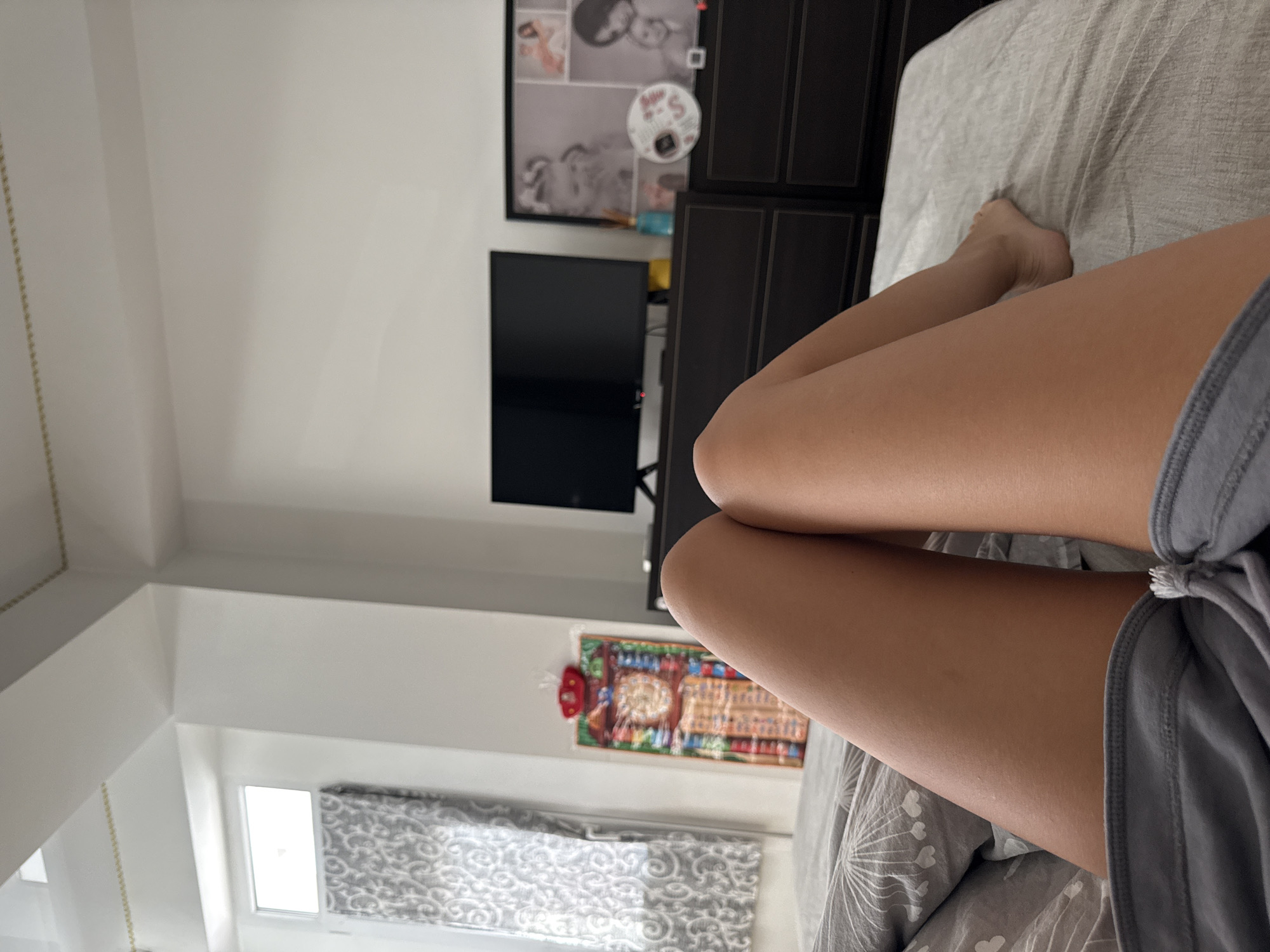 The image depicts a bedroom scene. In the foreground, a person's legs and feet are visible, resting on a grey bed with a patterned duvet. The person is wearing grey shorts. The background shows a dresser with a television set on top and framed pictures. To the left, a window with patterned curtains is visible. The overall environment appears to be a domestic setting, likely a bedroom.