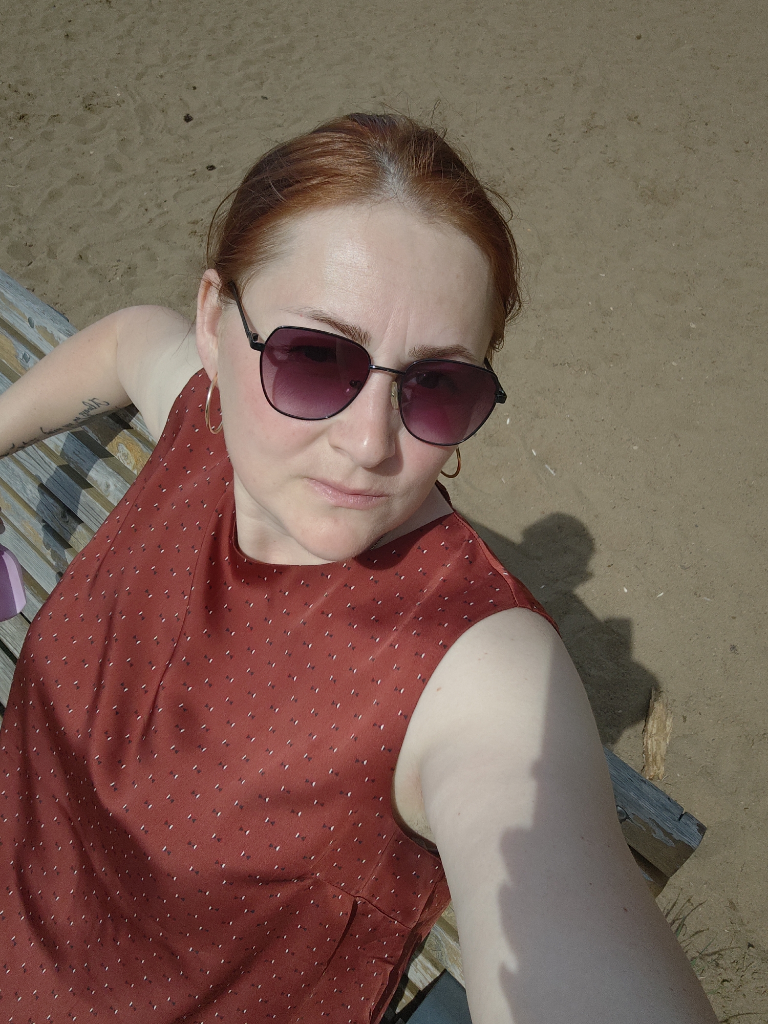 A red-haired woman wearing sunglasses and a sleeveless red dress with small white dots is sitting on a wooden bench. She is positioned at an angle to the camera, appearing to be taking a selfie. The bench appears to be outdoors, and the background shows sand. This photograph was taken in the city of Chaikovsky, Russia.