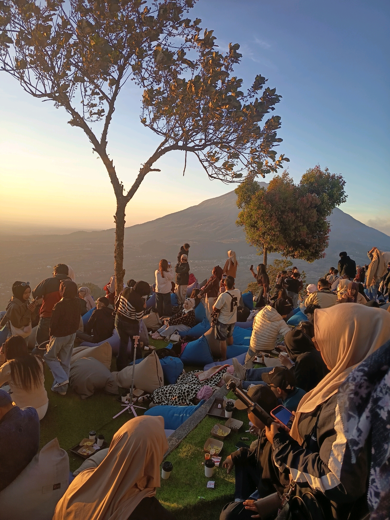 The image depicts a gathering of people on a grassy hillside overlooking a valley and a mountain range, likely in the city of Kediritan, Indonesia, during sunset. The scene is characterized by a relaxed atmosphere, with many individuals seated on large beanbag chairs scattered across the terrain.
In the foreground, a group of people are casually interacting, some holding phones, and others engaged in conversation. A young woman in the lower right is prominently visible, wearing a light-colored hijab and a dark jacket, holding a smartphone. To her left, a person in a black hoodie and baseball cap is also looking at a phone. A tray with food items, including French fries and cups, is placed on the grass nearby.
Further into the scene, more people are spread out on beanbags, enjoying the view. Some are standing and taking pictures, while others are seated, possibly observing the sunset. A solitary tree with sparse foliage stands in the mid-ground, with a larger tree to its left. In the background, a majestic mountain dominates the horizon, bathed in the warm glow of the setting sun, with a hazy valley stretching out below. The sky is a clear blue, transitioning to a warm yellow and orange hue near the horizon, indicative of twilight.
The overall impression is that of an outdoor social gathering or event, possibly a scenic viewpoint or a festival, where people have congregated to enjoy the natural beauty of the landscape at dusk.