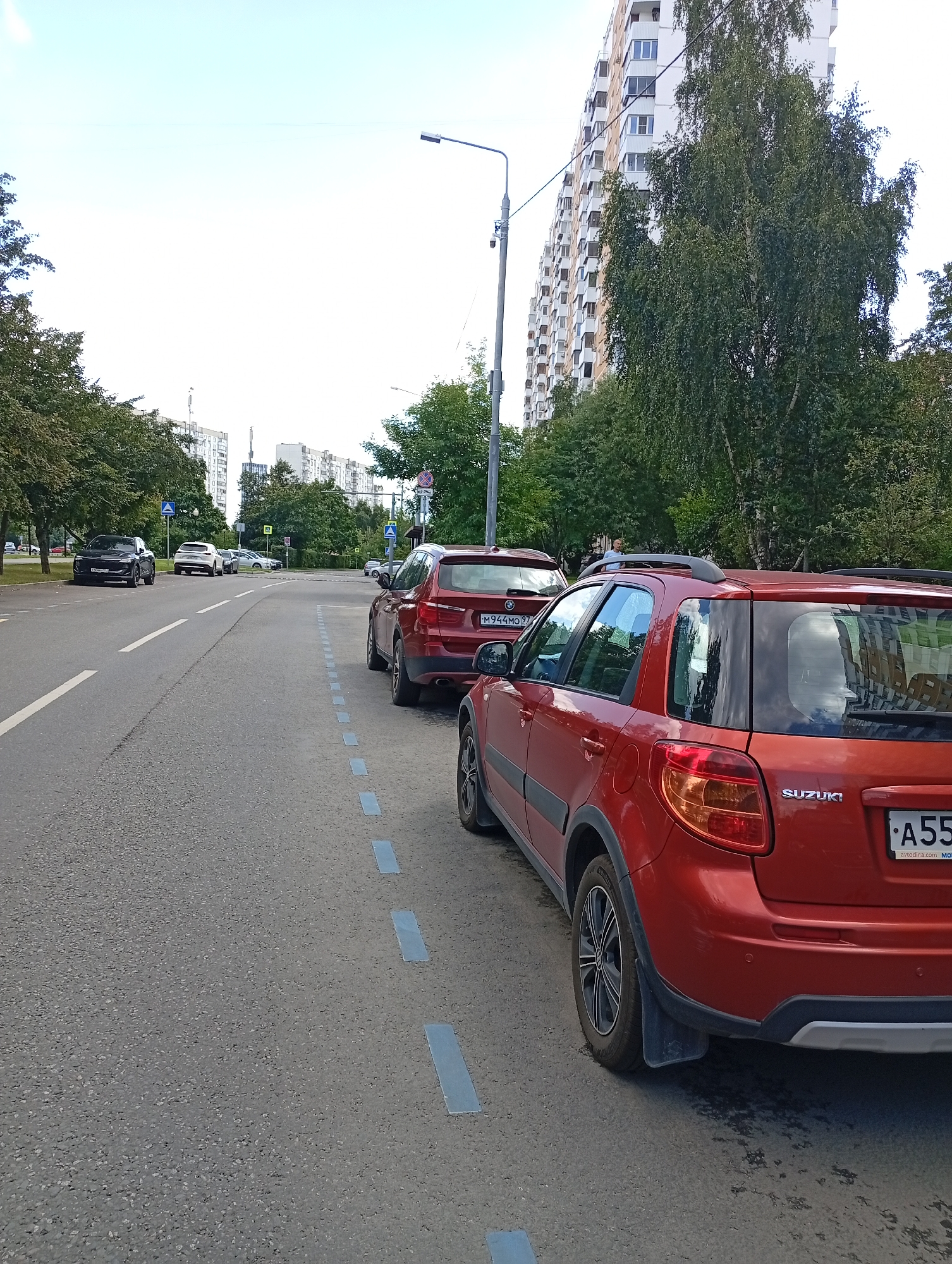 The image captures a street scene in an urban environment. In the foreground, a red Suzuki SX4 is parked, facing away from the camera. The car has a license plate that is partially visible, reading "A55". To the left of the Suzuki, another red car is parked, with a license plate that reads "M944MO 97". Further down the street, a black SUV is parked on the left side of the road. In the background, tall residential buildings line the street, with trees and a street light visible. The sky is overcast. The road surface appears to be asphalt, with blue painted markings indicating parking spaces.