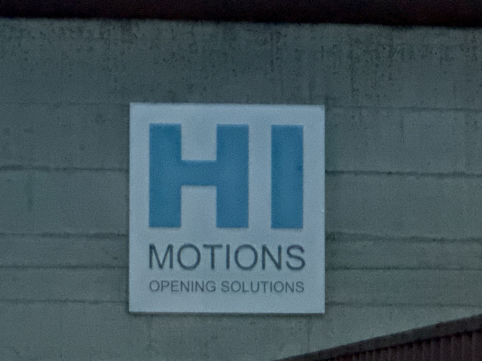 A rectangular white sign is affixed to a textured, light greenish-grey wall in Sarcedo, Italy. The sign features two large, light blue, uppercase sans-serif letters "HI" at the top. Below "HI," the word "MOTIONS" is rendered in smaller, dark grey uppercase sans-serif text. Below "MOTIONS," the phrase "OPENING SOLUTIONS" is displayed in even smaller dark grey uppercase sans-serif text. The wall exhibits horizontal lines, consistent with siding or block construction. A dark, indistinct horizontal structural element is visible along the top edge of the image, and a dark, vertically striated horizontal structure is present along the bottom edge. No individuals are present.