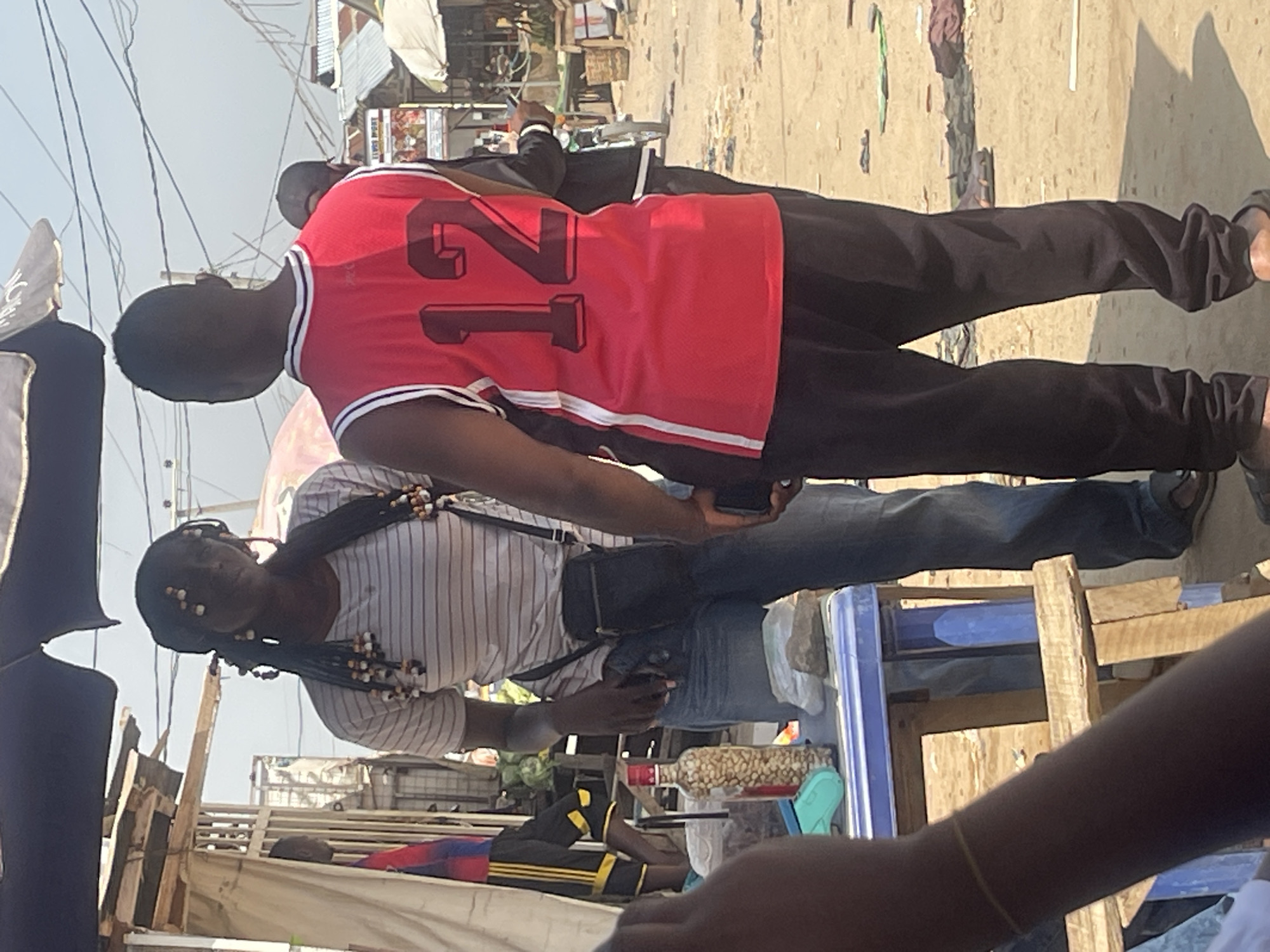 Two individuals, a woman and a man, are standing outdoors in what appears to be a marketplace in Abuja, Nigeria. The woman, positioned on the left and facing left, has long, braided dark hair adorned with beads. She is wearing a horizontally striped t-shirt and blue jeans, with a black crossbody bag slung across her body. In front of her is a blue plastic table displaying a glass bottle filled with nuts, a teal electronic device, and other items. The man, on the right and facing away from the camera, is wearing a red sleeveless jersey with the number "12" prominently displayed in black, and black pants. His back is to the viewer. In the background, various market stalls, some with corrugated metal roofs, are visible, along with power lines overhead and other people and vehicles in the distance. The ground is dusty and uneven, indicative of an unpaved area.