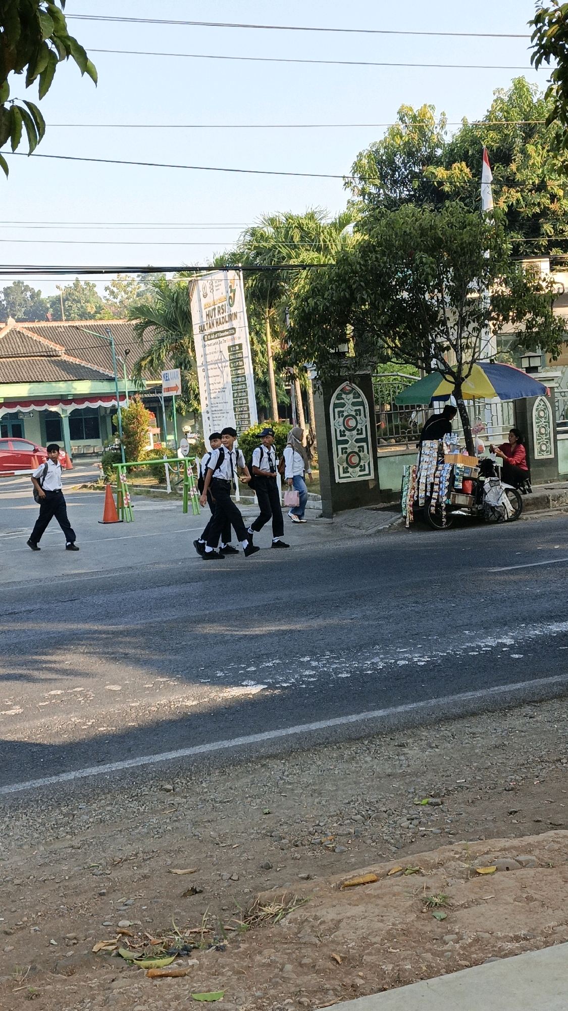 In Kuwasen, Indonesia, a street scene unfolds on a sunny day. A group of four uniformed schoolboys, wearing white shirts and dark trousers, are crossing a paved road. One boy walks ahead on the left side of the road, while the other three walk together in a line across the pedestrian crossing. They are accompanied by a girl in a white headscarf and a light blue shirt, carrying a pink bag. On the right side of the road, a woman in a red shirt sits behind a mobile vendor cart laden with snacks and beverages, shaded by a colorful umbrella. A motorcycle is parked next to the cart. In the background, a building with red and white trim is visible, along with trees and a white banner with text in Indonesian. The street has a white line marking a lane and a gravel shoulder.