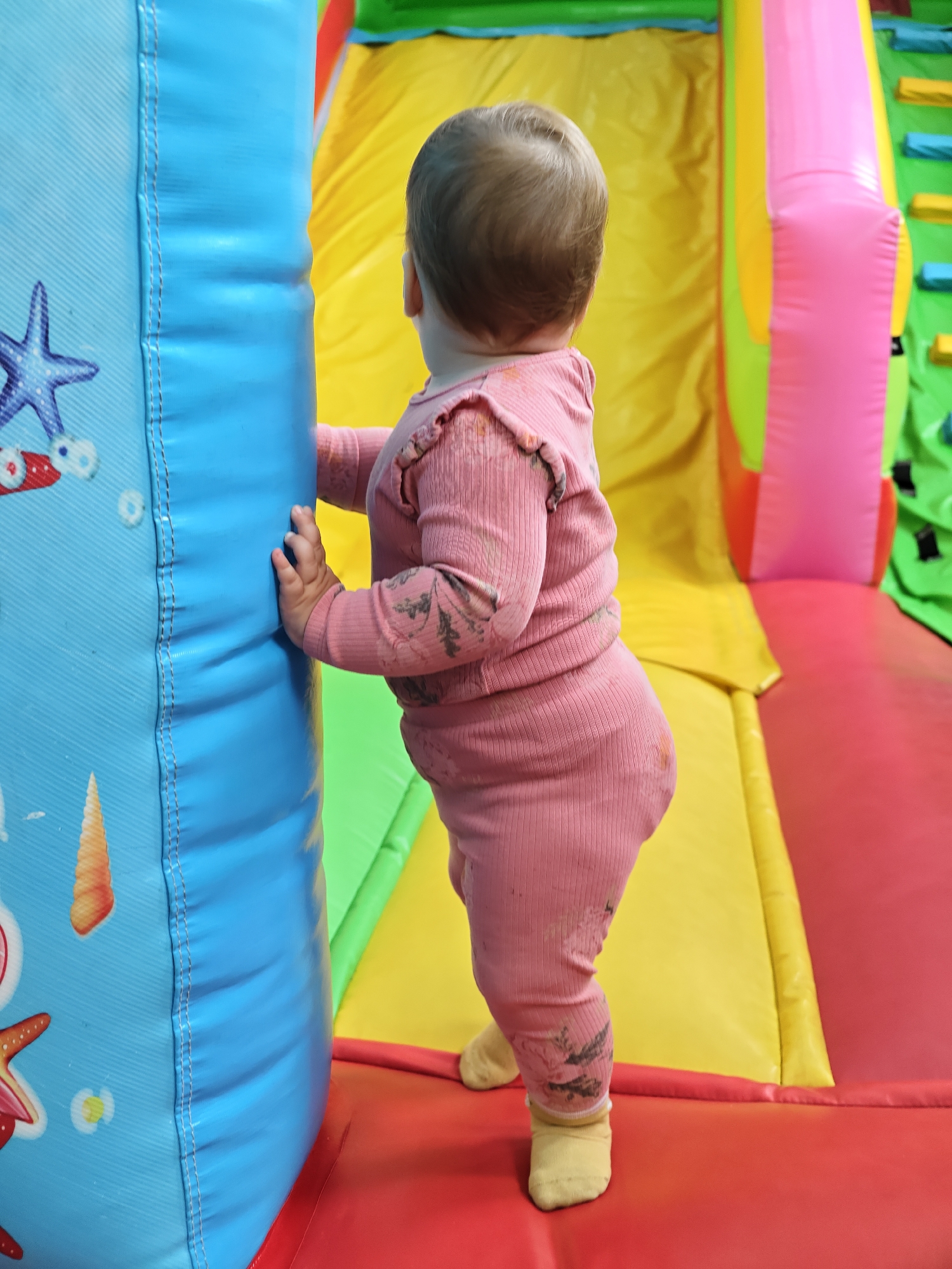 An infant is positioned in an indoor inflatable play area within the city of Chaikovsky, Russia. The child, viewed from behind, has light brown hair and is wearing a long-sleeved, ribbed, pink garment with subtle dark patterns, along with yellow socks. The child stands upright, supporting itself by grasping a vertical, blue inflatable pillar with its right hand. This blue pillar features printed maritime motifs, including a partially visible purple starfish and an orange conch shell, and displays prominent vertical stitching. The play area's surface is a red inflatable mat. In the background, behind the child, a multi-colored inflatable structure is visible, featuring a large yellow section, a bright pink vertical inflatable element, and sections of green inflatable material that appear to be steps or climbing aids. The child is facing the yellow and pink sections of the play structure.