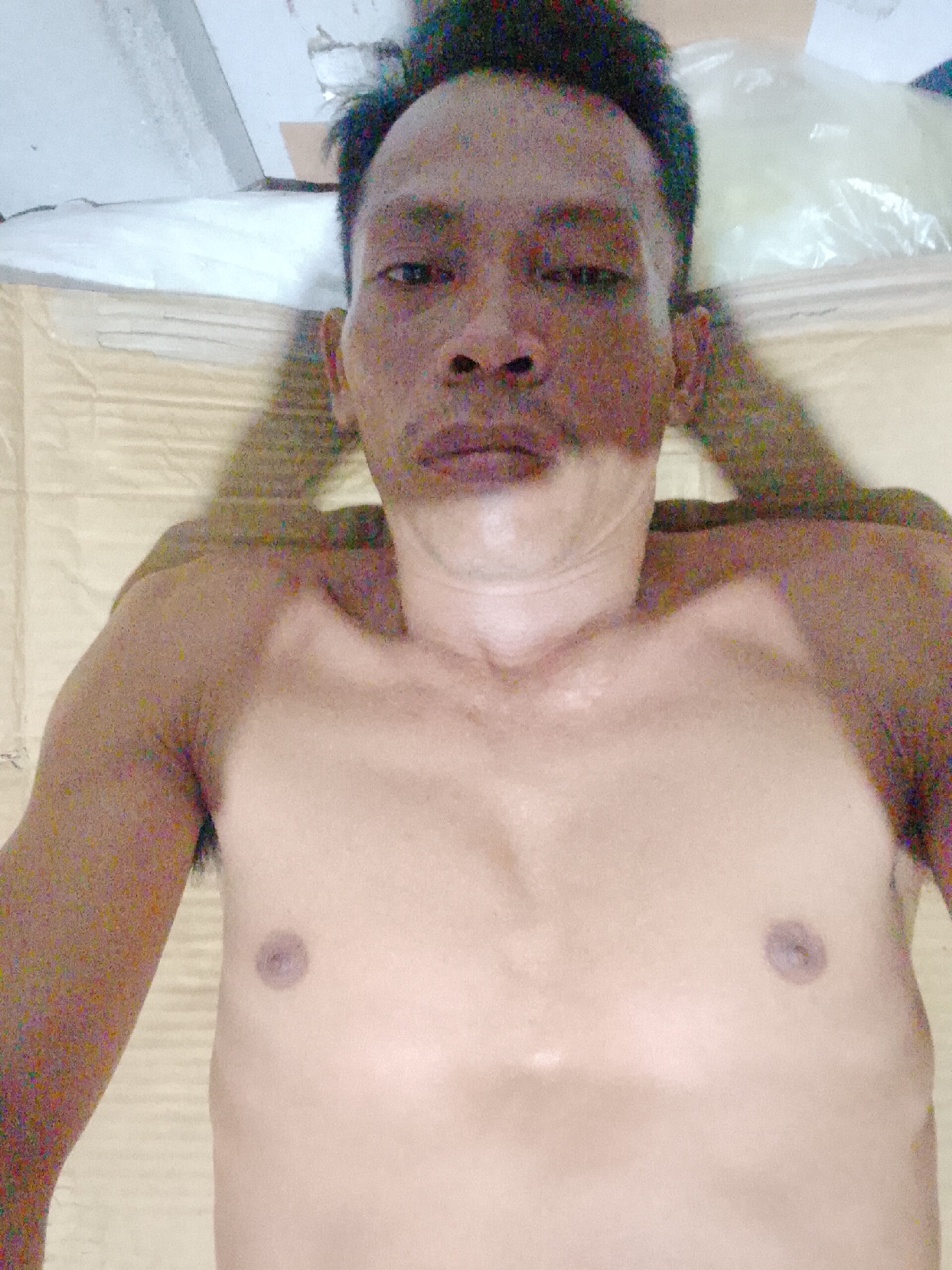 The image shows a shirtless man lying on what appears to be a padded surface. The location, Bukit Ketok, Indonesia, is not discernible from the visual information. The man has short, dark hair and a mustache. He is looking directly at the camera. His arms are positioned near his shoulders. The environment is indistinct but shows a white ceiling in the background, along with what appears to be plastic packaging material.