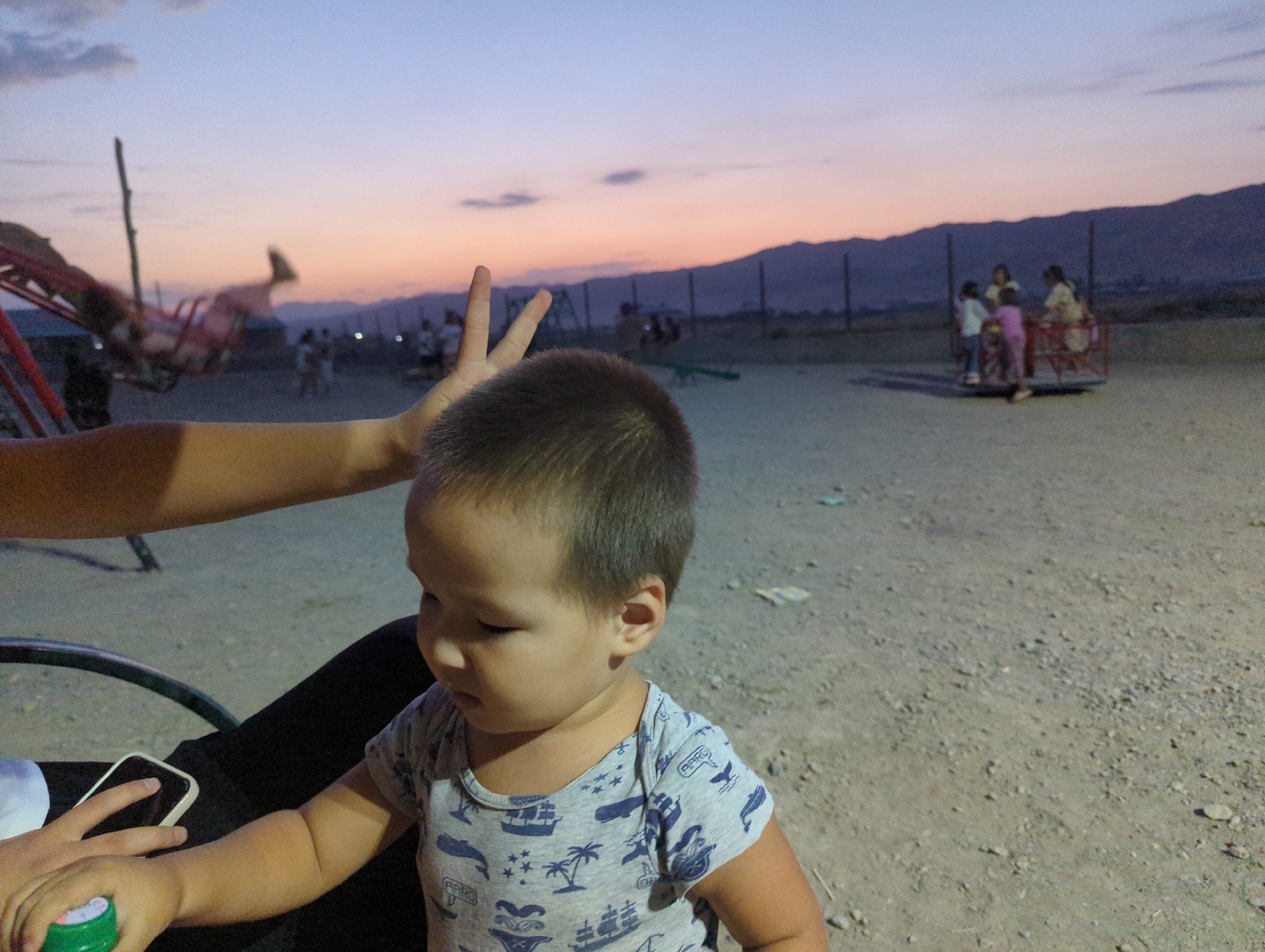 At dusk in a dusty outdoor area, possibly a playground or park in the village of Uch-Dobo, Kyrgyzstan, a young child in a grey onesie with a nautical print is the central focus. The child is looking downwards, seemingly engaged with something out of frame. In the foreground, an adult's arm, which is holding a smartphone and a small green object, is visible near the child. In the background, a blurred merry-go-round with several figures, likely children and adults, is present. A swing set structure and mountains are also visible in the distance under a twilight sky.