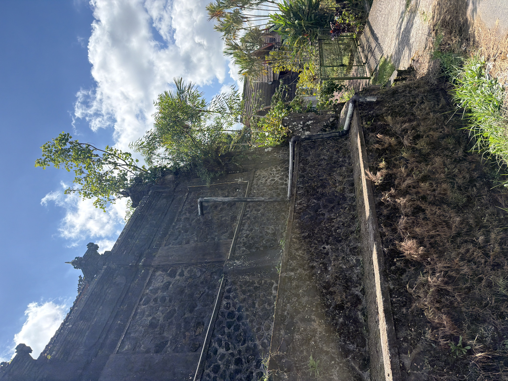 The image depicts a view of an old stone wall and surrounding vegetation in Baturiti, Indonesia. The wall, constructed with rough-hewn stones and some smoother cemented sections, exhibits signs of age and weathering, with moss and vines growing on its surfaces. Two visible pipes run along the wall, one horizontal and one vertical, suggesting some form of drainage or utility. Beyond the wall, a glimpse of tropical foliage, including palm trees, and a traditional Indonesian building with a tiled roof are visible. A metal gate and a paved pathway leading to the property are also present. The sky above is bright blue with scattered white clouds, indicating a clear day. The scene appears to be located in a rural or semi-rural setting within Baturiti. No people are present in the image.