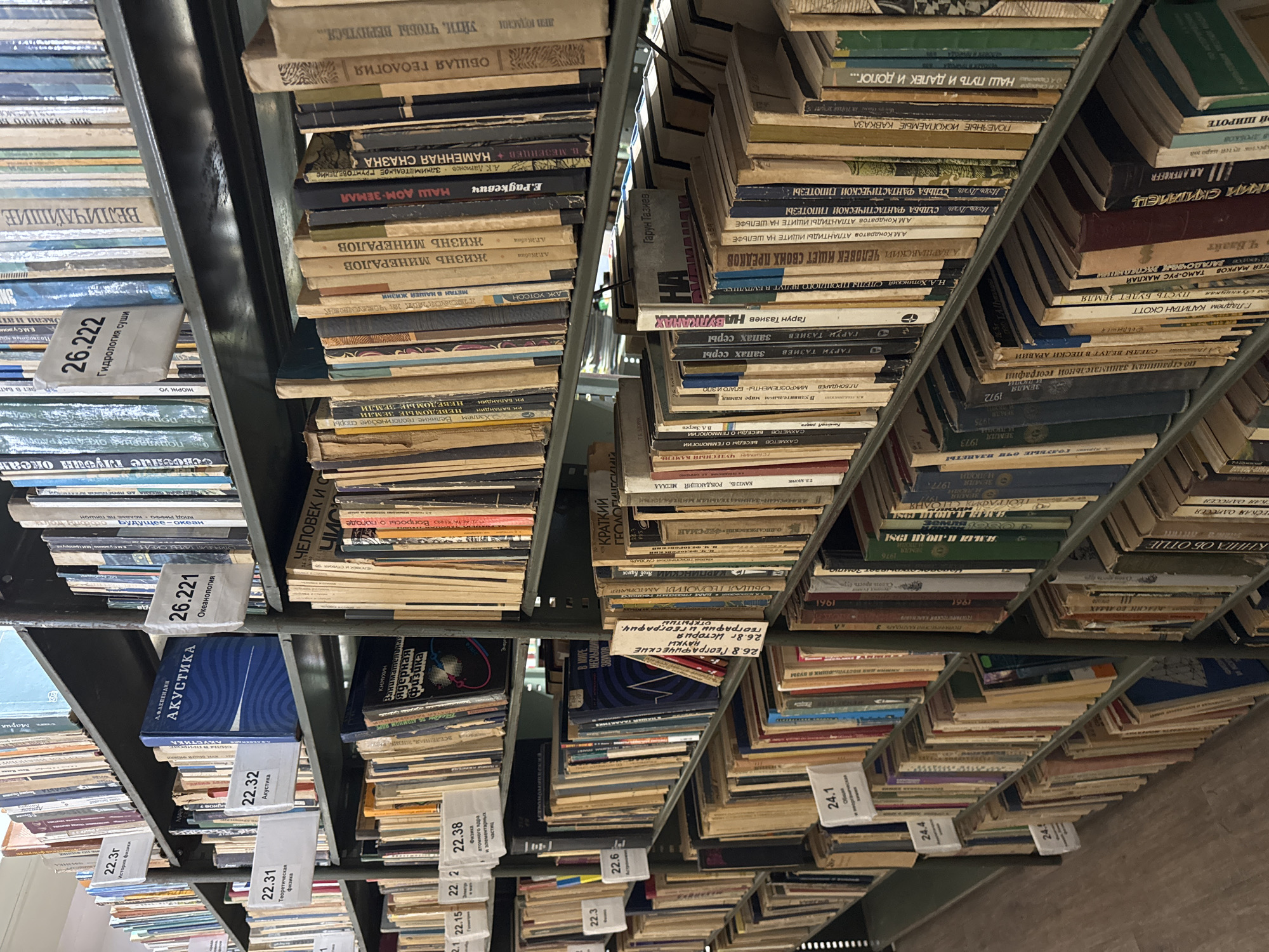 The image shows a library shelf filled with books. The books are of various sizes and colors, and some have labels with numbers and titles. The shelves are made of metal, and the lighting suggests an indoor environment. The overall impression is one of an organized and well-stocked library.