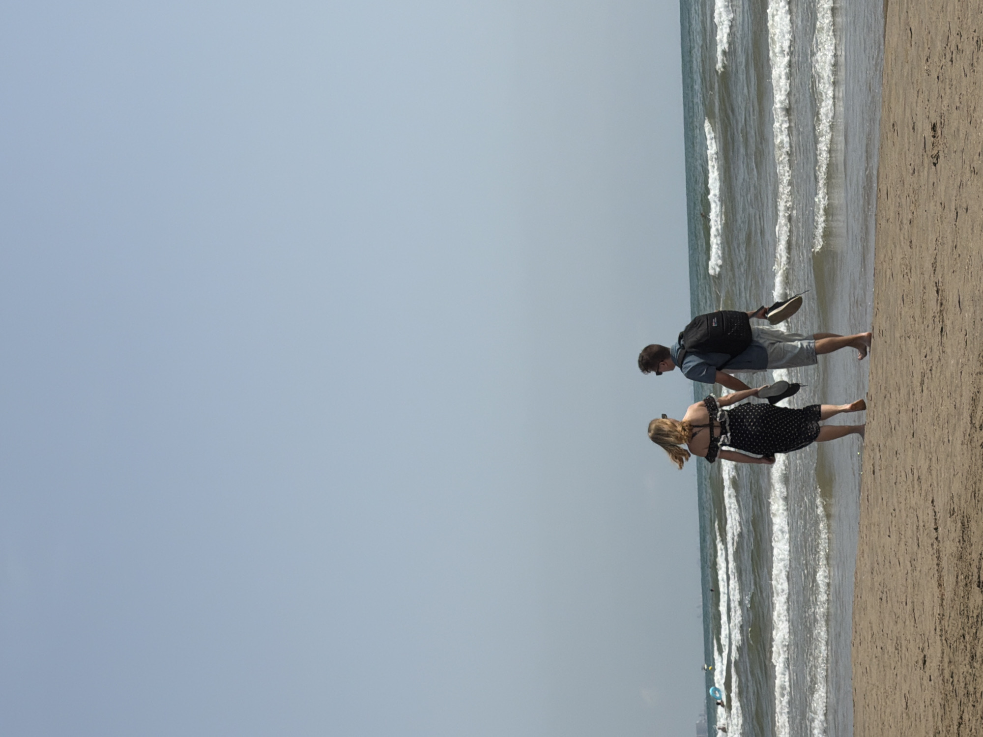 A rear view of a couple walks along a sandy beach. The woman, on the left, wears a black and white polka dot dress and holds a pair of flip-flops in her right hand. The man, on the right, wears a blue short-sleeved shirt, grey shorts, and a black backpack. He also holds a pair of flip-flops in his right hand. The ocean has white-capped waves and the sky is a clear, light blue. In the distance, a faint city skyline is visible.