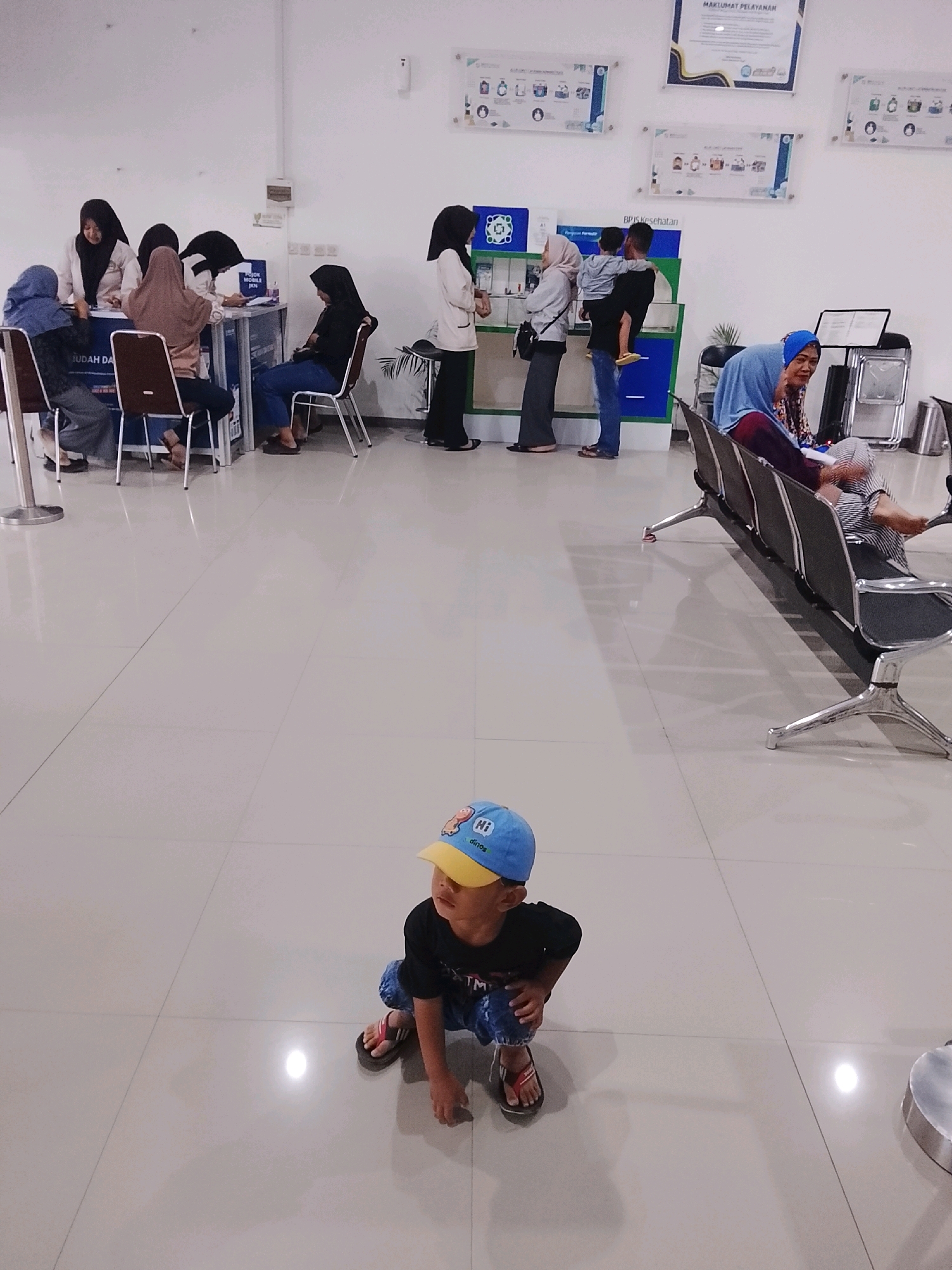 In a waiting area, likely a government office or clinic in Slawi, Indonesia, a young boy wearing a blue and yellow cap and a black t-shirt squats on a polished tile floor. He is facing away from the camera, his head tilted slightly to his right. To his left, a small circular reflection shines on the floor, possibly from a light source.
In the background, several people are seated or standing. To the left, a counter is staffed by women wearing hijabs, some of whom appear to be assisting clients. Other individuals are seated on a row of dark chairs with metal frames. Further back, a man holds a child, and a woman in a blue hijab is seated, looking towards the right. The walls are white, adorned with framed informational posters. The environment appears clean and organized, typical of a public service facility.