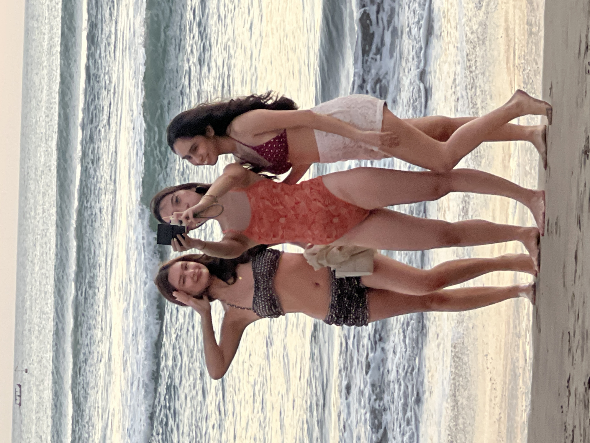 Three young women are posing for a selfie on the beach in Kuta, Indonesia. The woman on the left is wearing a patterned bikini set. The woman in the center is wearing an orange one-piece swimsuit and holding a camera. The woman on the right is wearing a polka dot bikini top and white lace shorts. The ocean is in the background with gentle waves. The sky is overcast and the light suggests it is either early morning or late afternoon. A small boat is visible in the distance on the horizon.