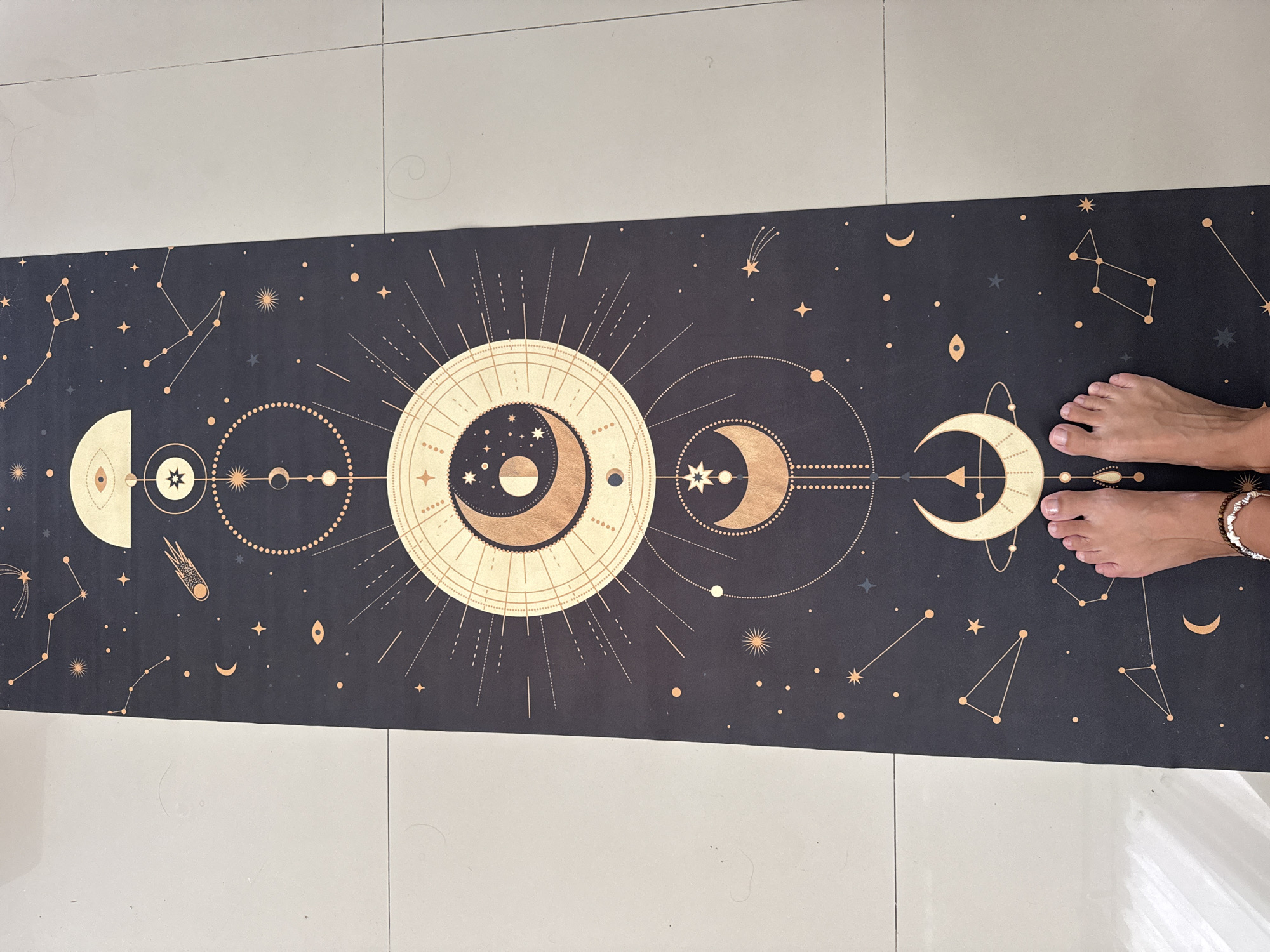 A yoga mat with a celestial design is lying on a tiled floor. The mat is primarily dark with gold-colored celestial bodies and constellations scattered across it. A pair of feet are positioned at the right end of the mat, wearing bracelets. The location is likely indoors in Kuta Selatan, Indonesia, given the tile flooring.
