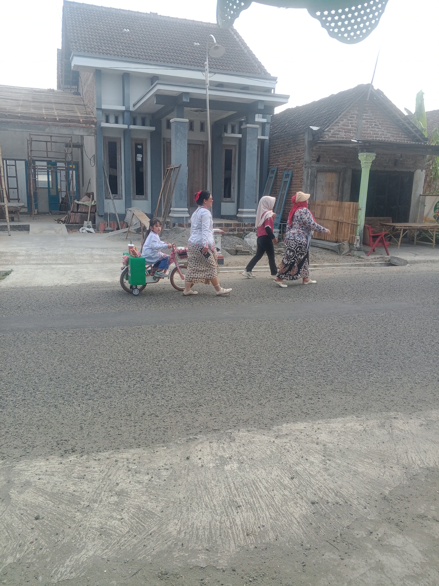 In Lembah, Indonesia, on a paved road, a child rides a pink toy bicycle with training wheels. The child, wearing a white shirt and blue pants, has a cup and a green container attached to the bicycle. A woman in a white shirt and patterned skirt walks beside the bicycle, holding a phone. Two other women, dressed in hijabs and modest clothing, walk in front of them on the same side of the road. One woman wears a pink hijab and a dark shirt with black pants, while the other wears a patterned dress and a red head covering. In the background, a light-colored house with dark trim and blue accents is under construction, with scaffolding and building materials visible. Adjacent to it, a brick building with a corrugated metal roof stands.