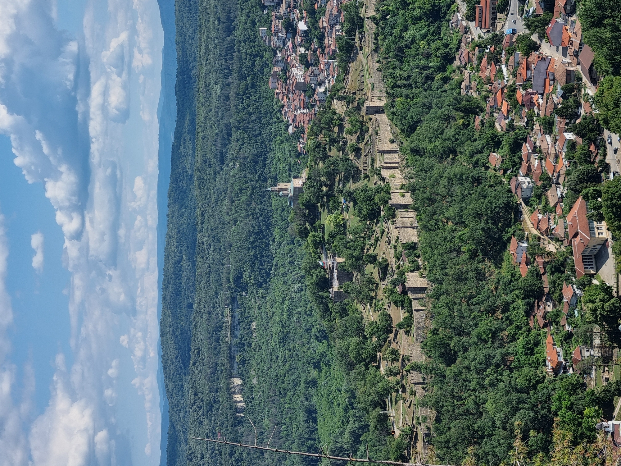 The image shows a panoramic view of the city of Veliko Tarnovo, Bulgaria, dominated by the Tsarevets fortress. The fortress, with its imposing stone walls and towers, is perched on a hill overlooking the city. A prominent church with a tall steeple stands within the fortress grounds. Below the fortress, a densely populated urban area with red-tiled roofs spreads across the landscape. The city is nestled amidst lush green hills and a dense forest. The sky is partly cloudy, with a mix of blue sky and white cumulus clouds. The overall scene depicts a historic city in a natural, scenic setting, likely captured on a bright, sunny day. The image is a high-angle shot, offering a broad perspective of the cityscape and its surroundings.